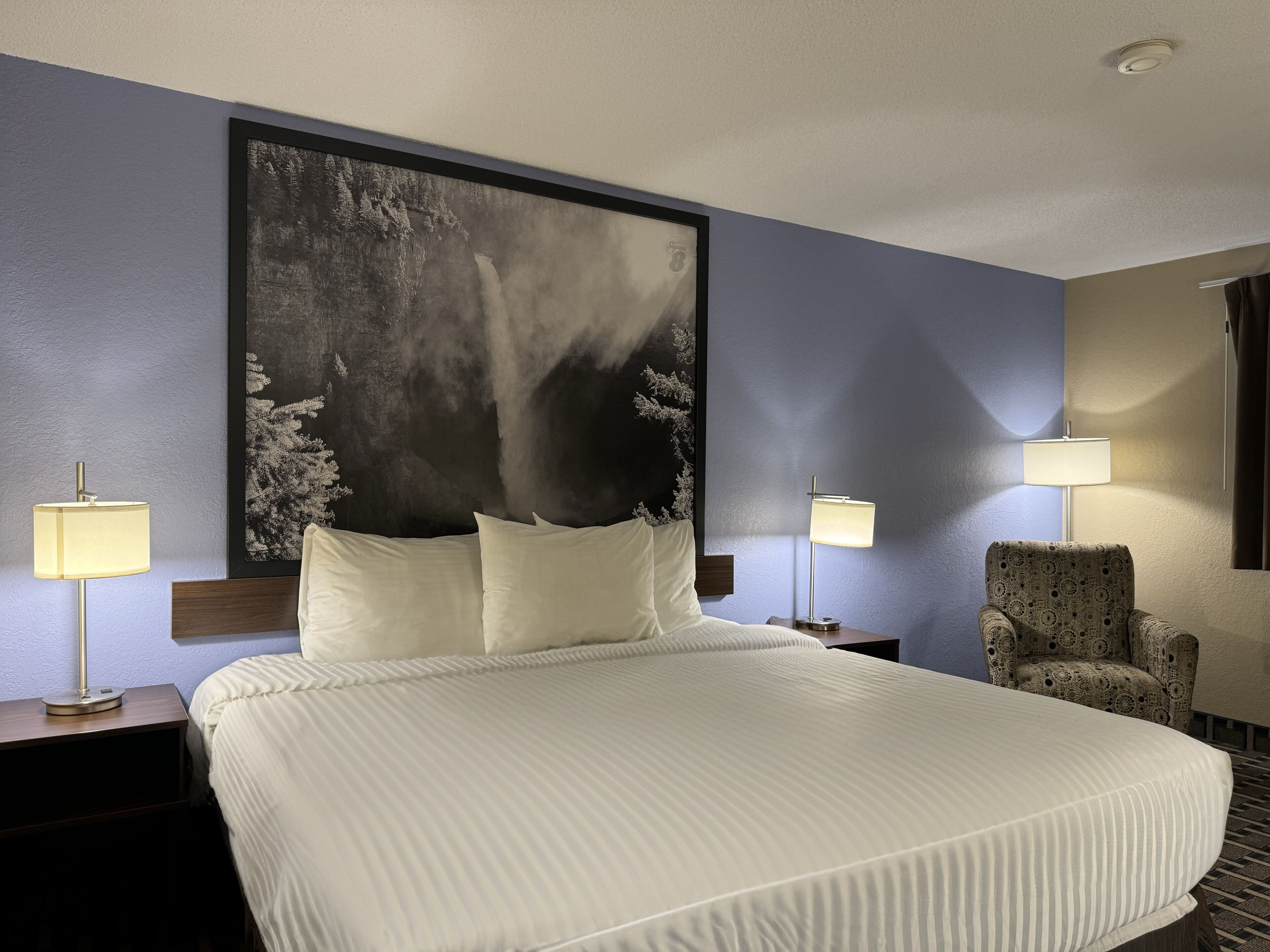 A hotel room with a large bed, two bedside lamps, a patterned armchair, and a large framed picture on the wall.