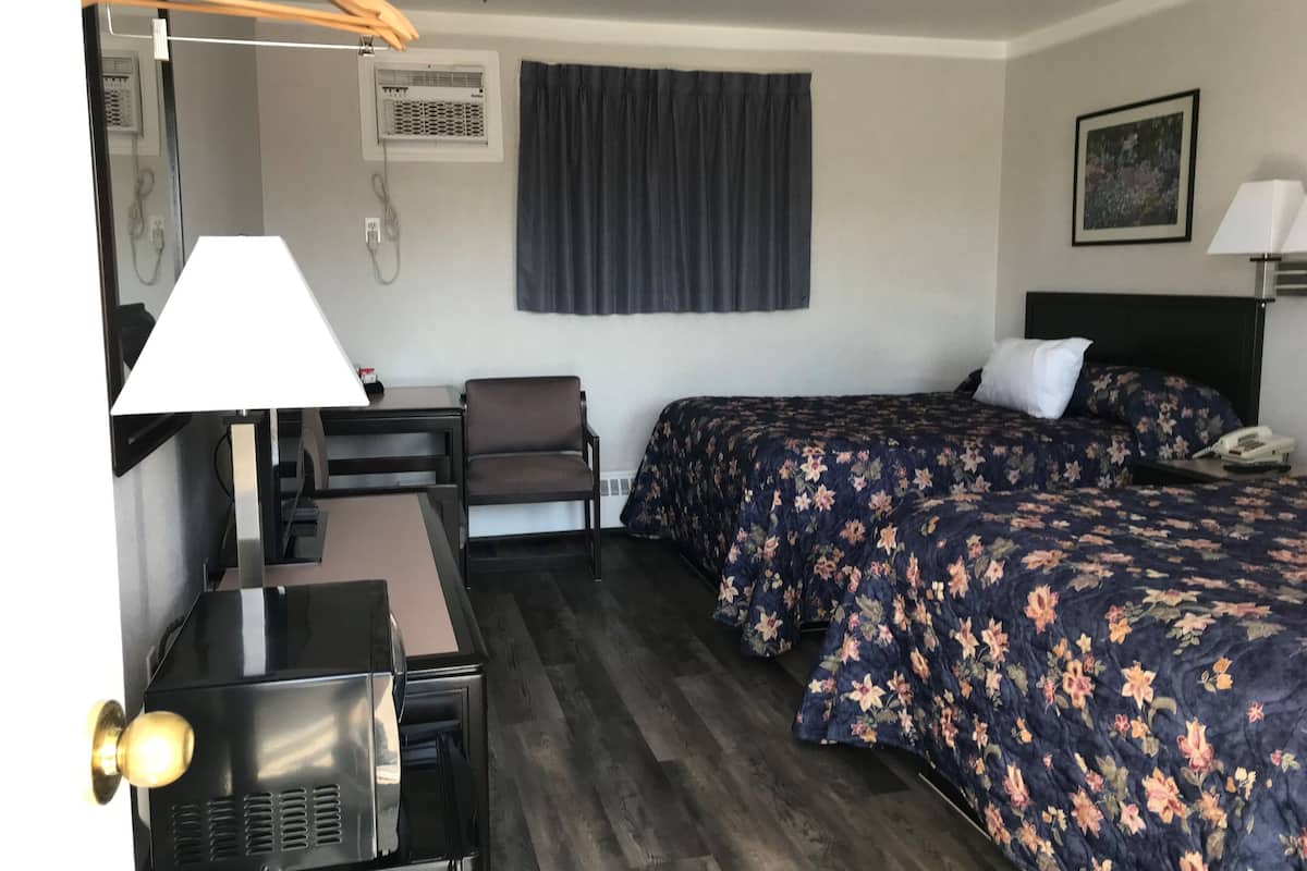 A hotel room with a desk, chair, two beds with floral bedding, a ceiling fan, an air conditioning unit, and a painting on the wall.