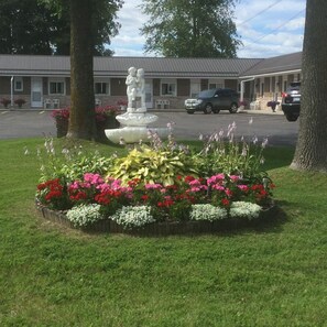 A landscaped garden with a fountain and colorful flowers, a motel building in the background, and a parked car.