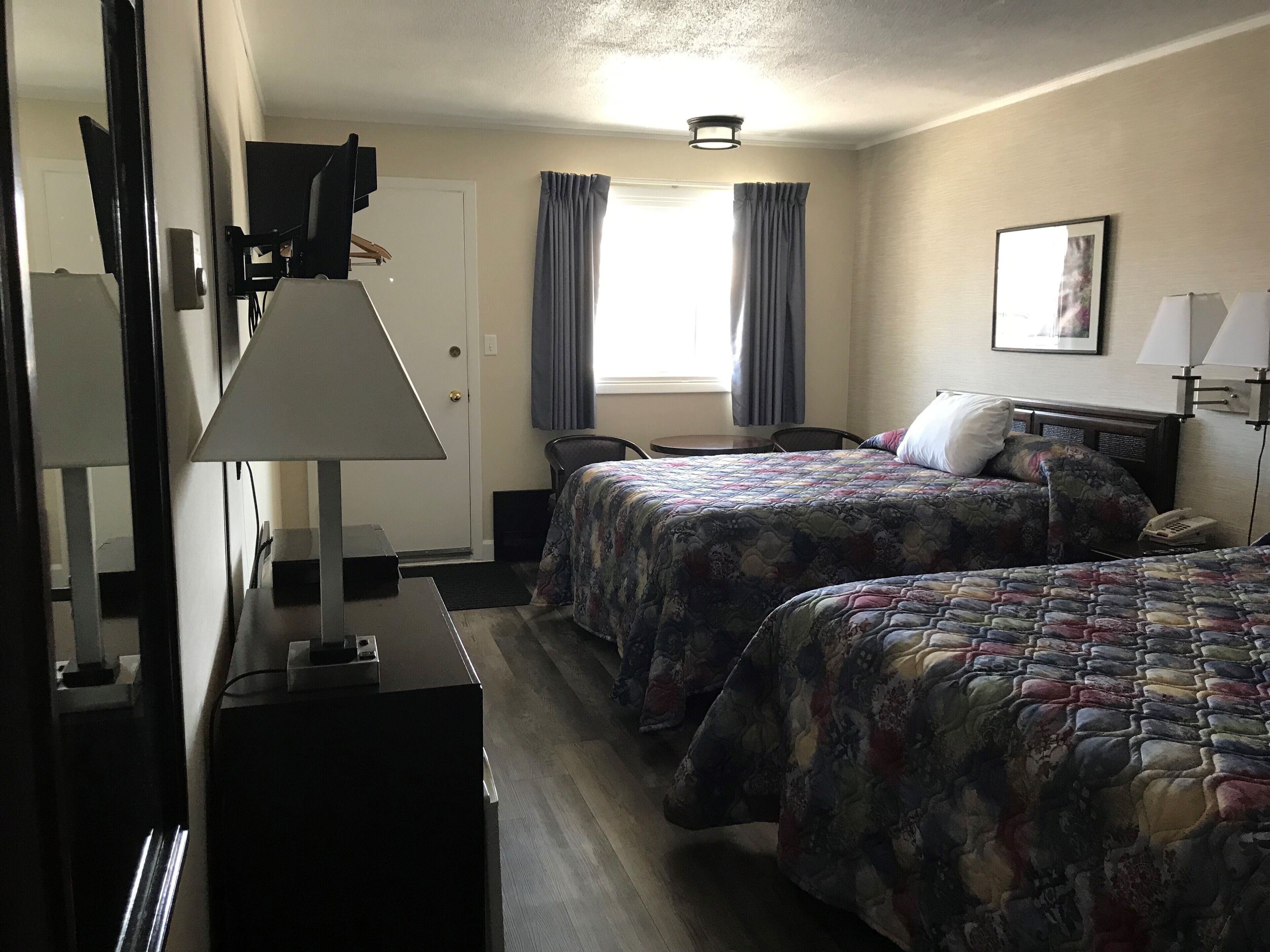 Habitación de hotel con dos camas, un escritorio, una silla, una lámpara, una ventana con cortinas y un cuadro en la pared.