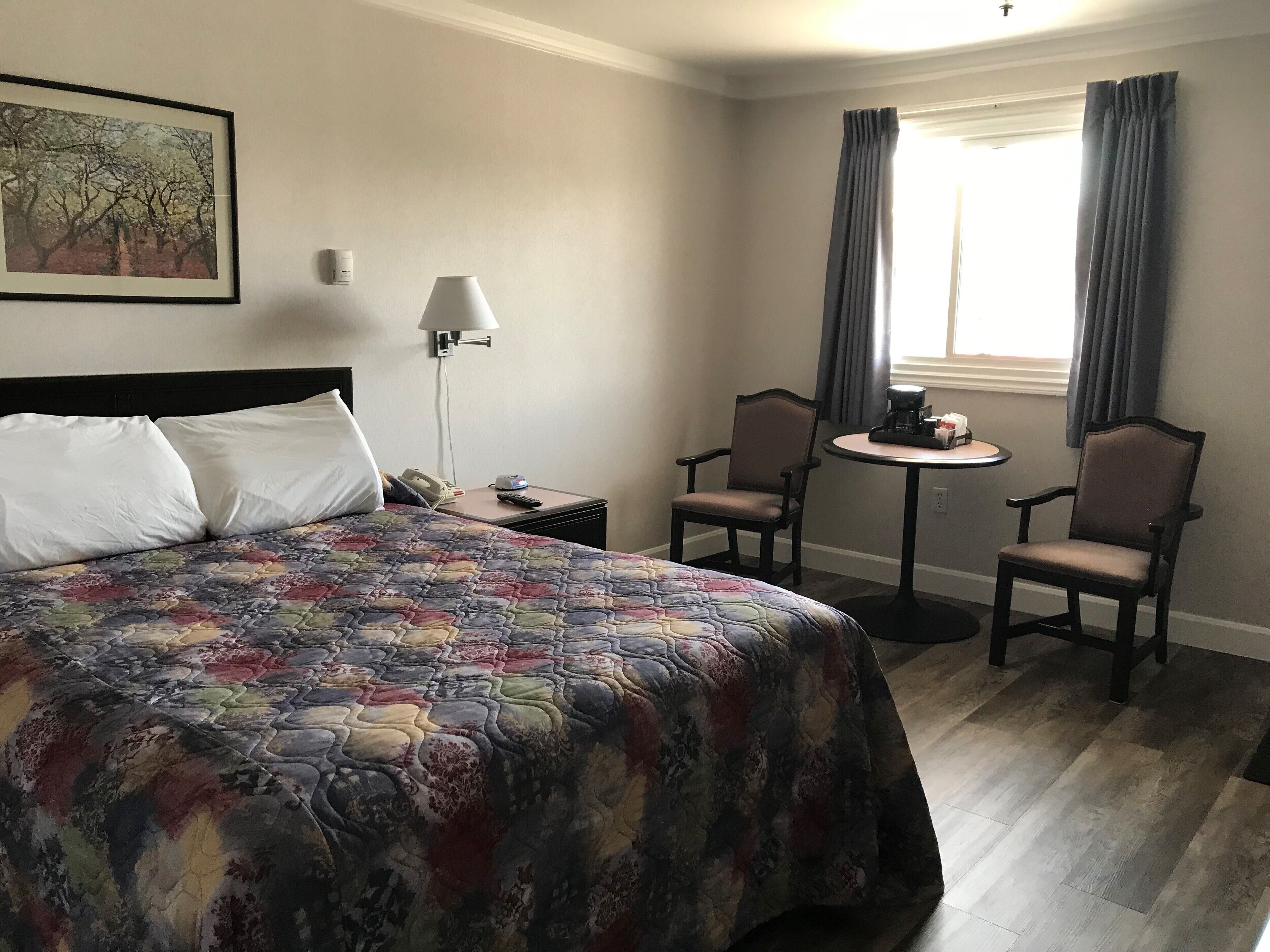 A hotel room with a large bed, two armchairs, a small round table, a lamp, a telephone, and a painting on the wall.