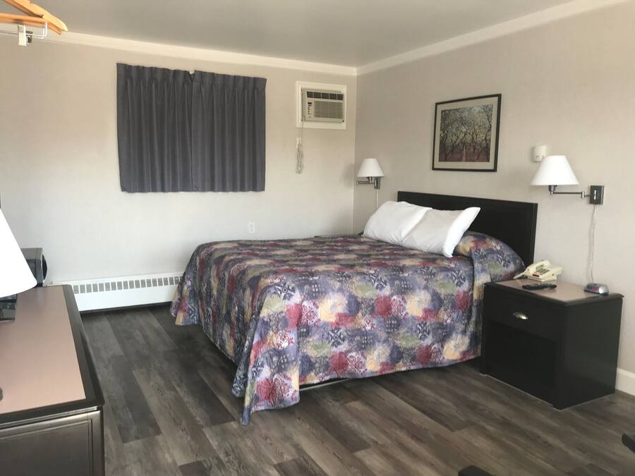 A hotel room with a bed, bedside lamps, a nightstand with a phone and remote, a desk with a lamp, and a window with curtains.