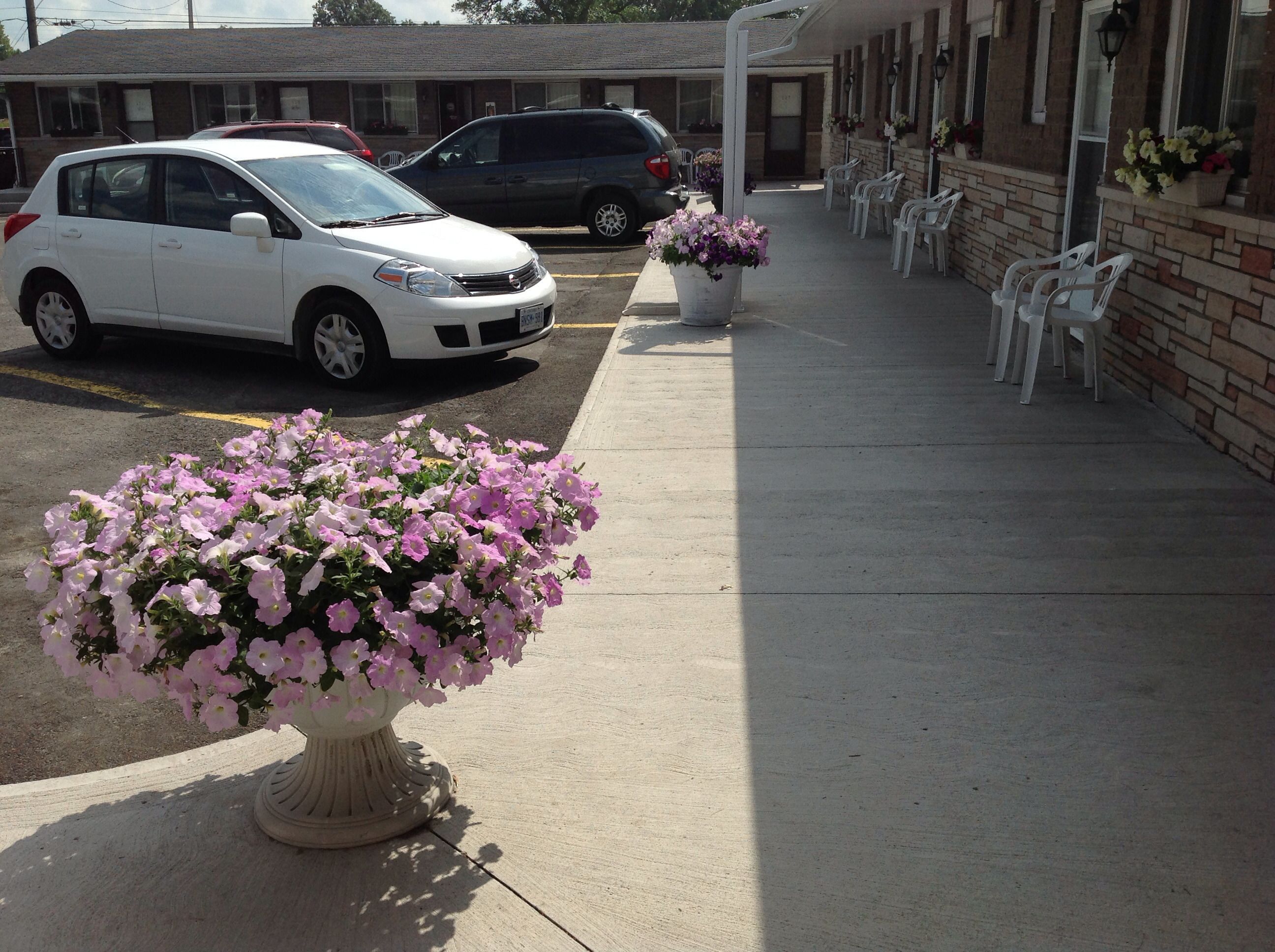 A parking area with a white car and a black SUV, a row of white plastic chairs, and a building with a brick facade.