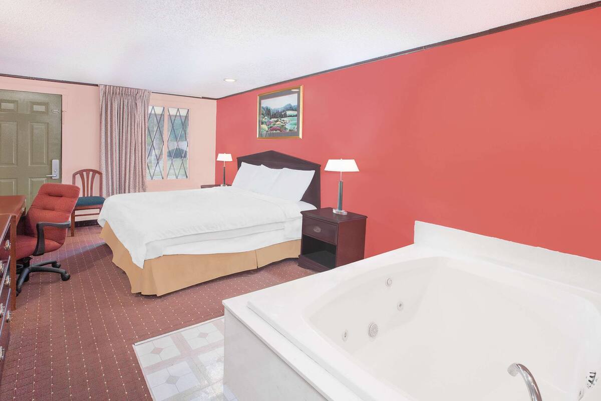A hotel room with a bed, a desk, a chair, a desk lamp, a bedside table, and a bathtub.