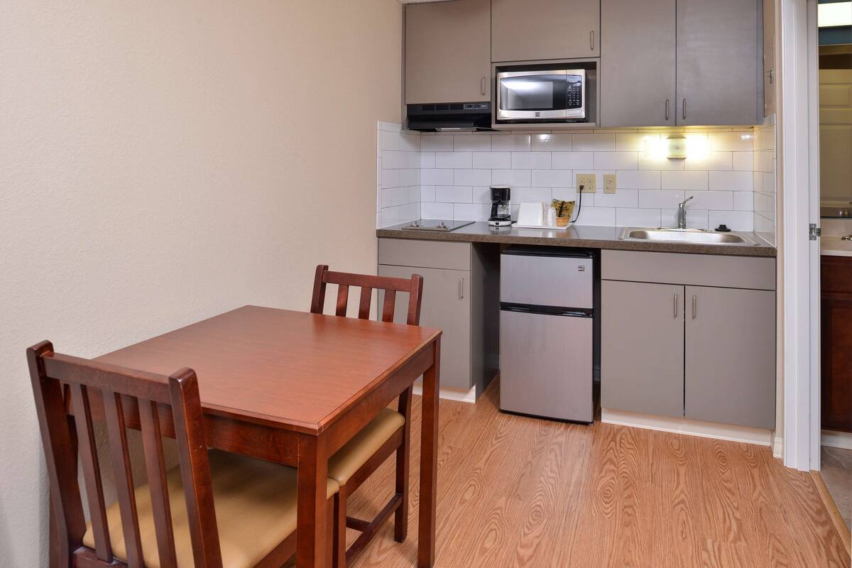 Cocina compacta con una mesa y sillas de madera, un microondas, una cafetera y un refrigerador.