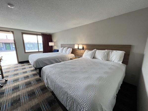 Quarto de hotel com duas camas, uma janela grande, uma escrivaninha e um abajur.