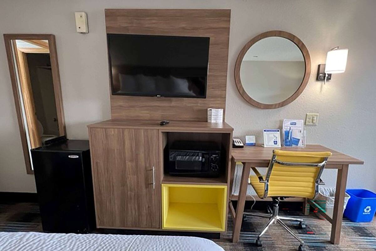 A hotel room with a bed, a TV mounted on a wooden cabinet, a desk with a chair, a mirror, and a trash bin.