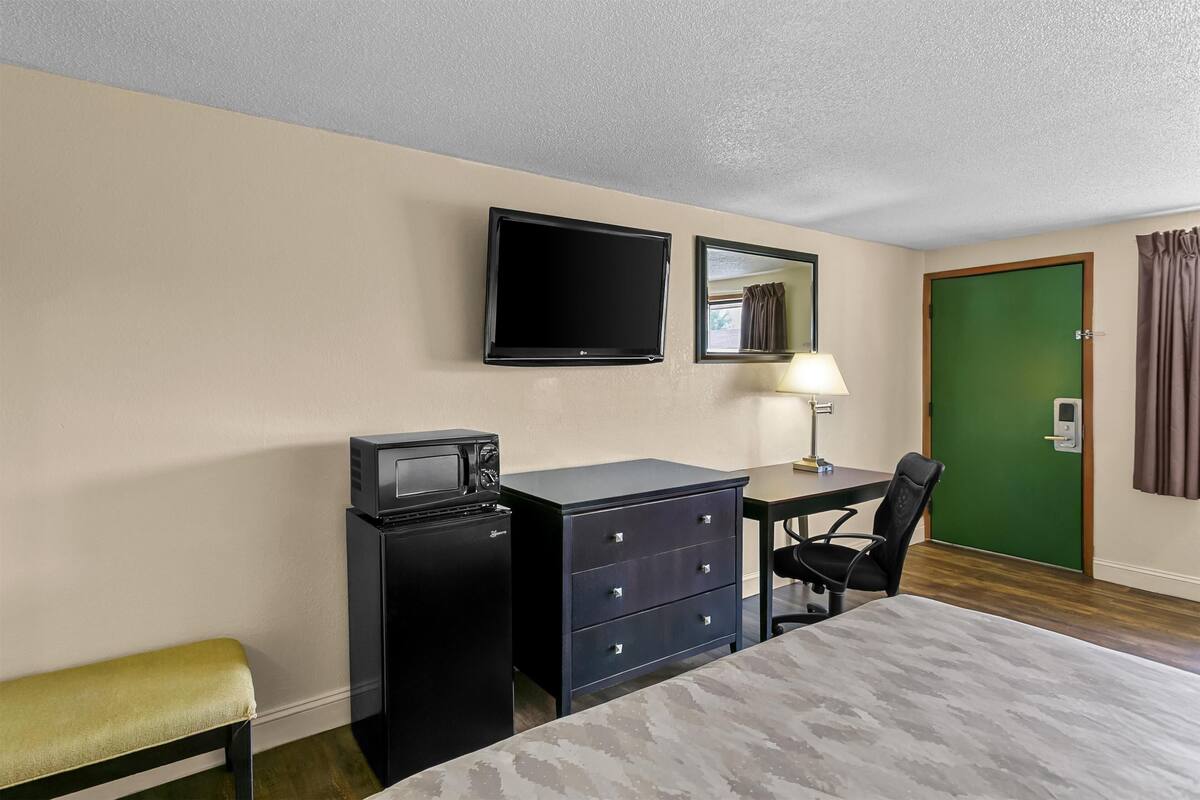 Una habitación de hotel con una cama, un escritorio con silla, un televisor, un microondas, un aparador y una puerta verde.