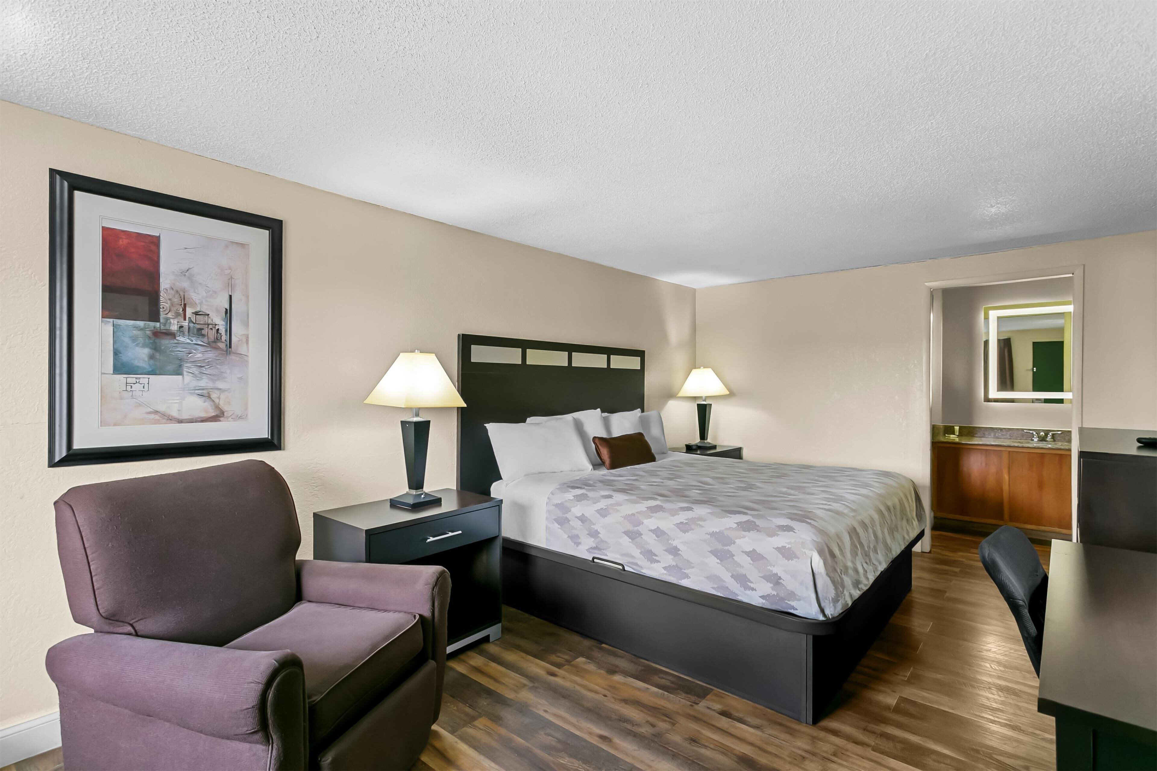 A hotel room with a bed, two bedside tables with lamps, a chair, a desk, and a framed picture on the wall.