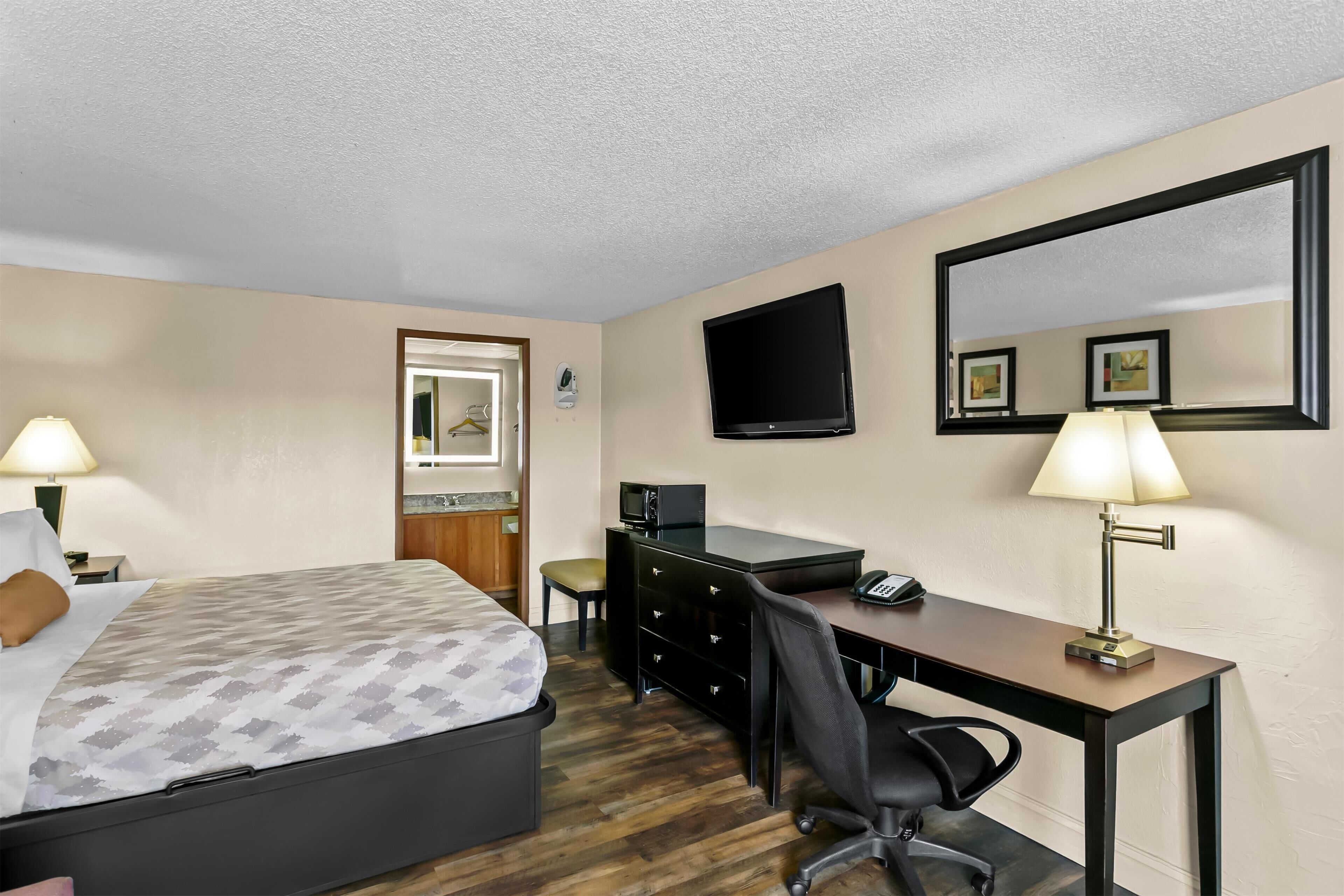 A hotel room with a bed, desk, chair, desk lamp, TV, and a door.