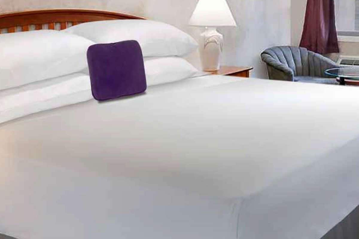 A neatly made bed with white linens and a purple pillow, a wooden headboard, a bedside table with a lamp, and a chair with a round table beside the bed.