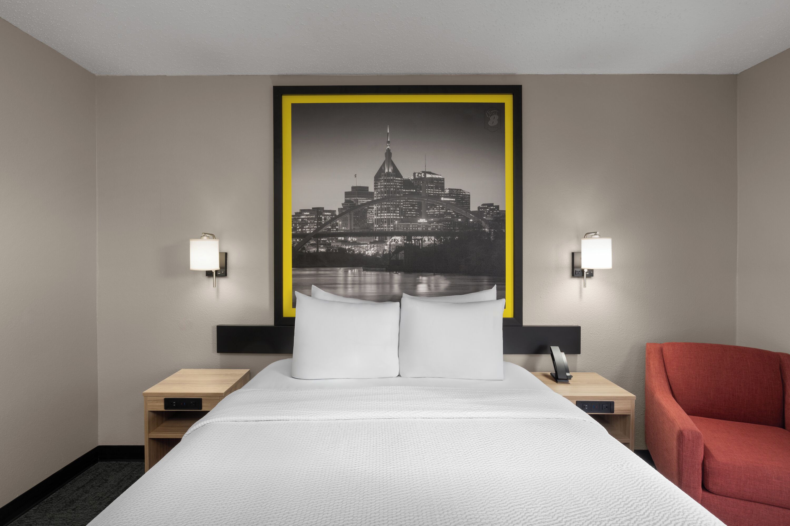 Quarto de hotel com uma cama, duas mesas de cabeceira, uma cadeira vermelha e uma obra de arte com vista para a cidade na parede.