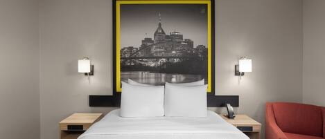 A hotel room with a bed, two bedside tables, a red chair, and a cityscape artwork on the wall.