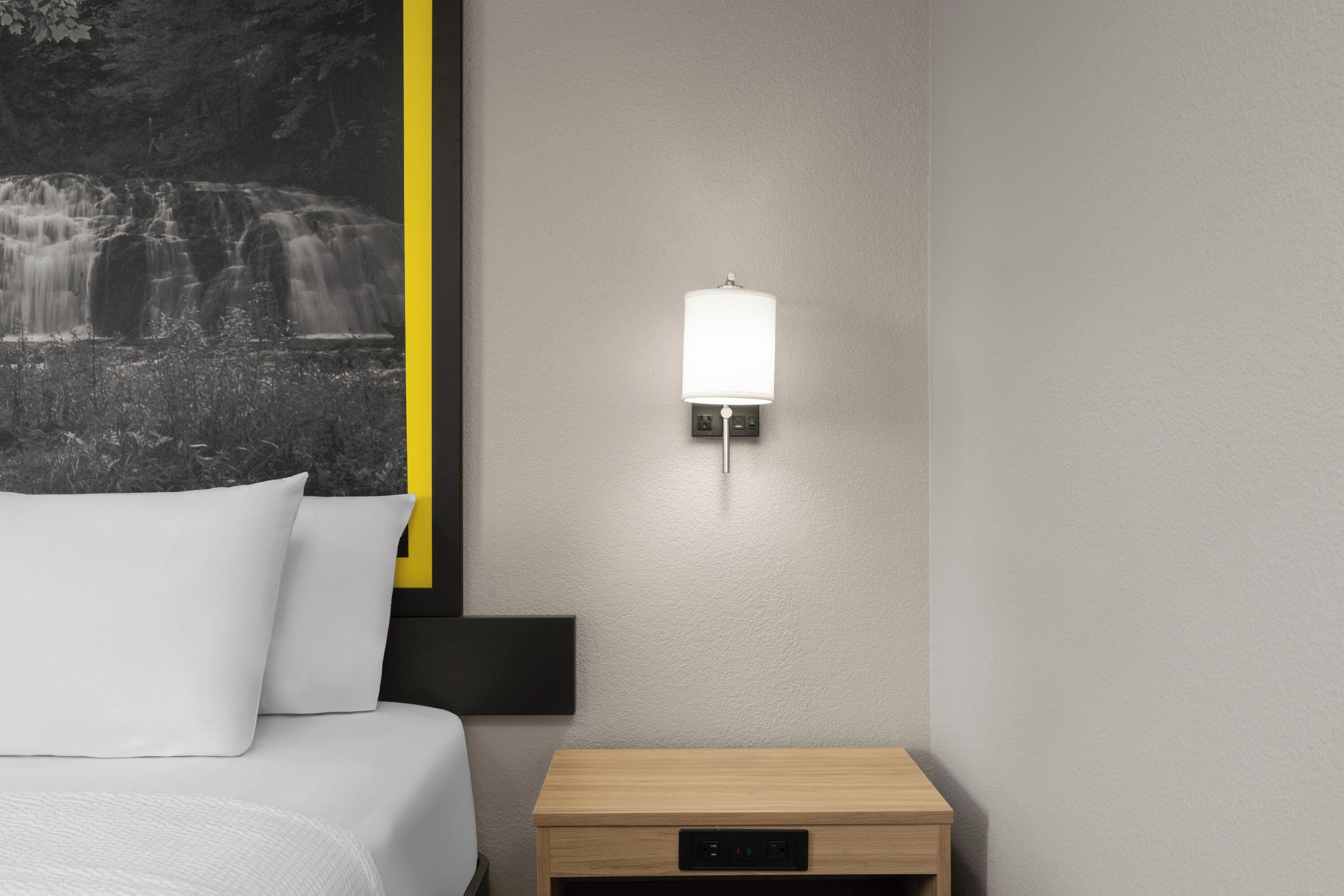 A bedroom with a bed, a nightstand, a wall-mounted lamp, and a framed picture of a waterfall.