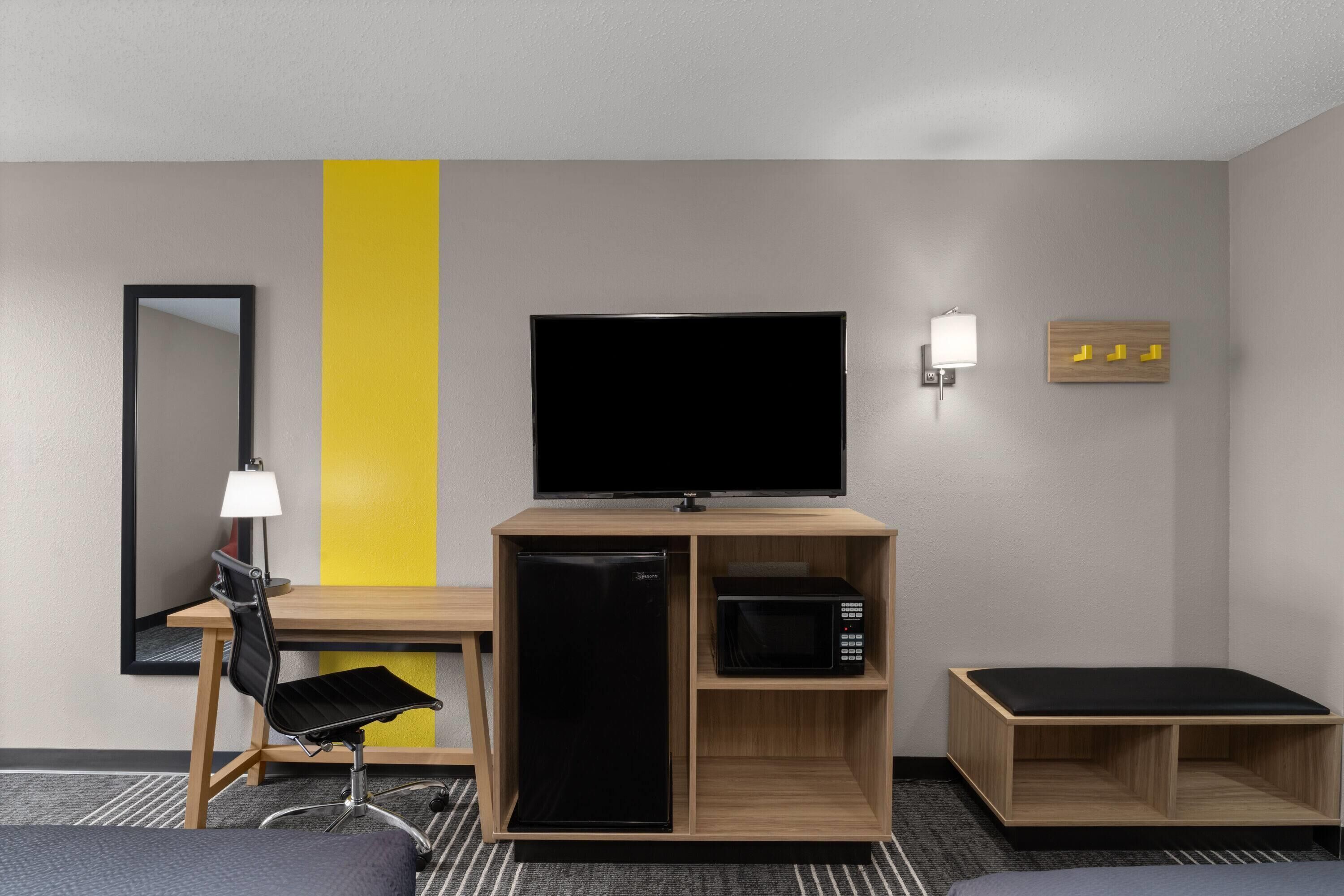 Habitación de hotel con escritorio, silla, televisión, refrigerador, microondas y una pequeña zona de estar.