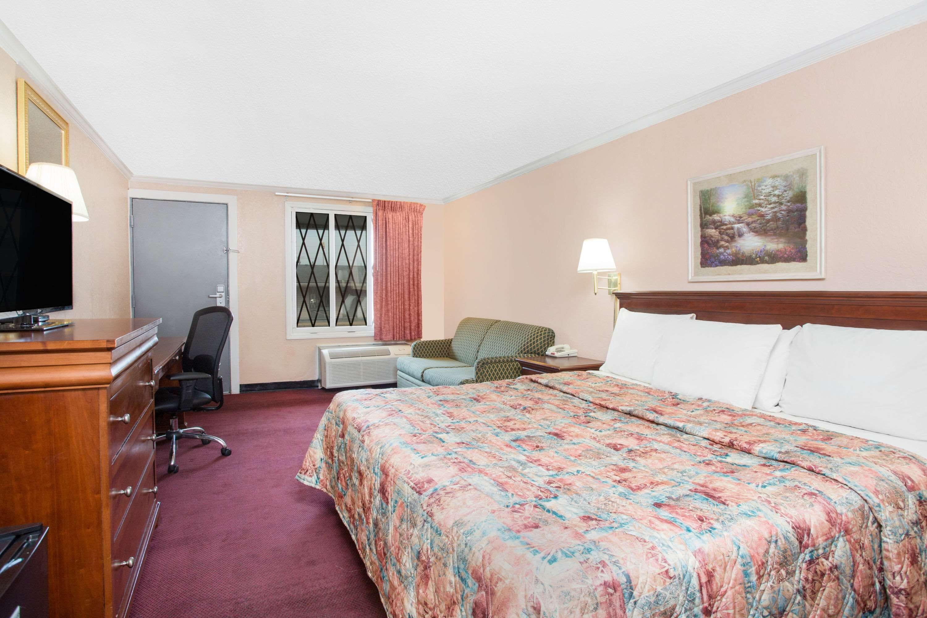 Een hotelkamer met een bed, een bureau, een stoel, een televisie, een raam met gordijnen en een schilderij aan de muur.