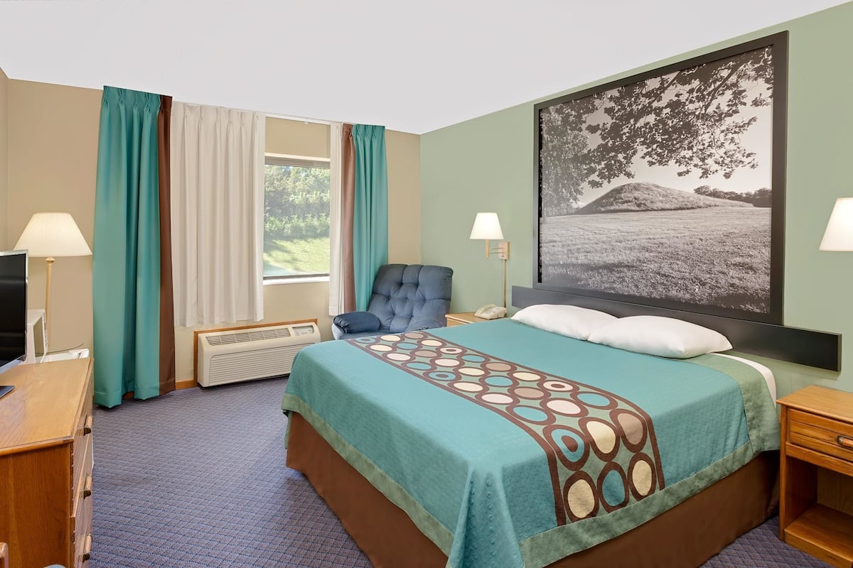 Quarto de hotel com uma cama, uma cadeira, uma escrivaninha com televisão, um abajur, um aquecedor e um quadro grande emoldurado na parede.