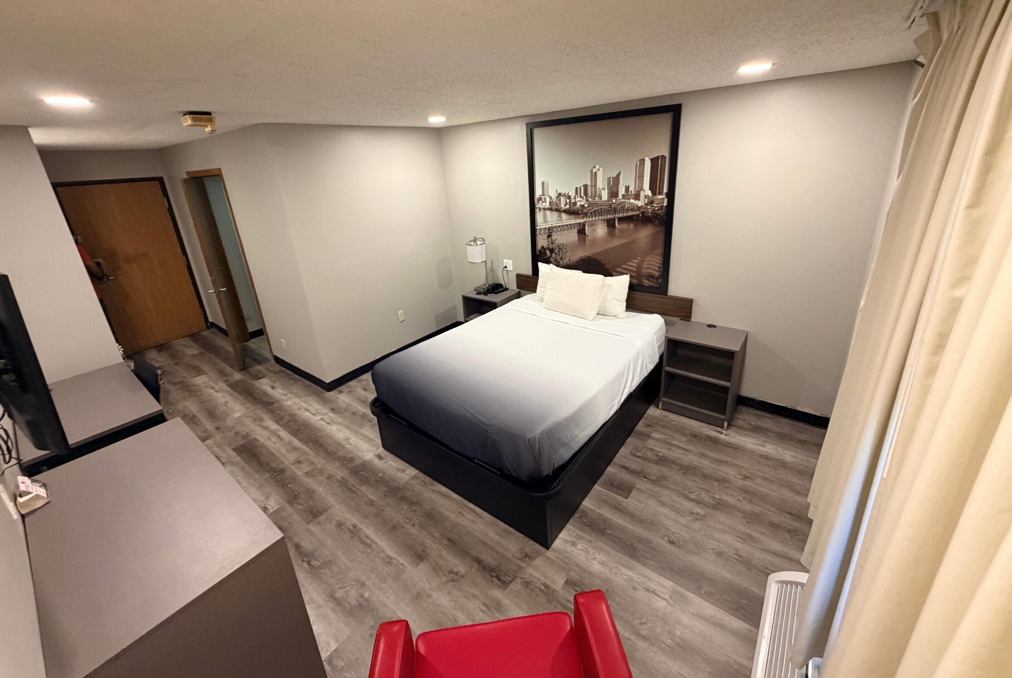 A hotel room with a bed, bedside table, lamp, and a cityscape painting on the wall.