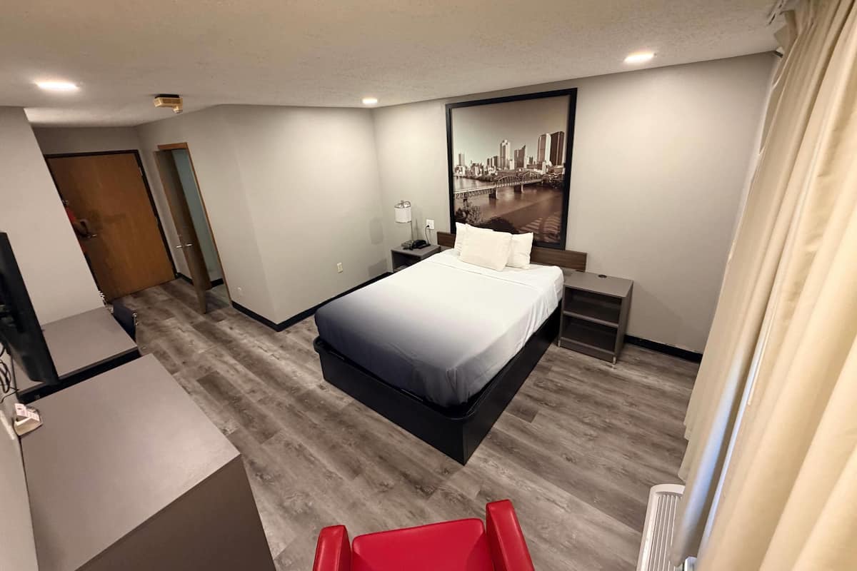 Quarto de hotel com cama, mesa de cabeceira, abajur e uma pintura da paisagem urbana na parede.