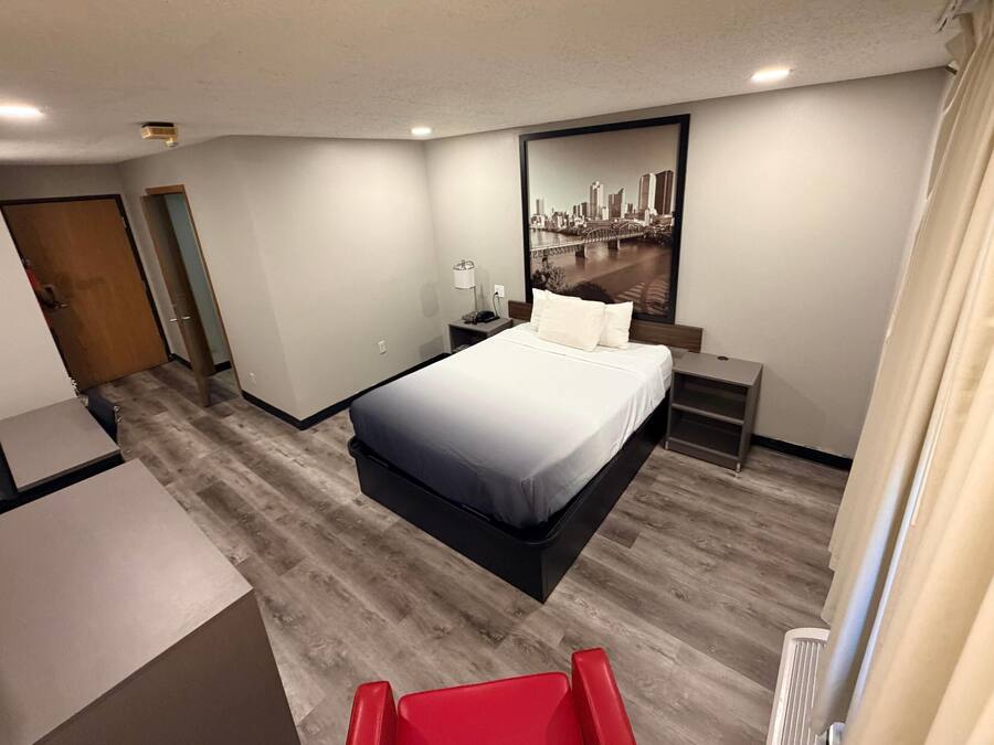 A hotel room with a bed, bedside table, lamp, and a cityscape painting on the wall.