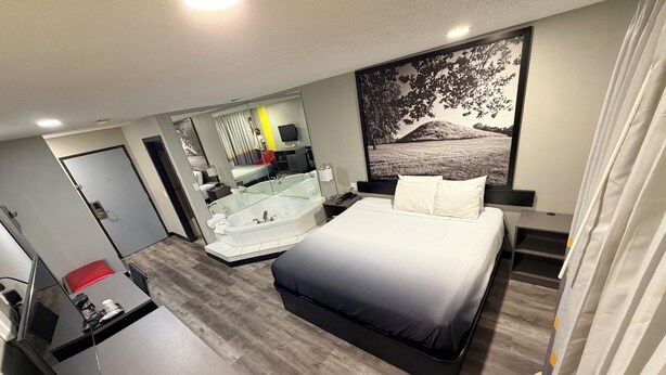Une chambre d’hôtel moderne avec un grand lit, un bureau, une chaise rouge, une baignoire et une grande fenêtre donnant sur un paysage.