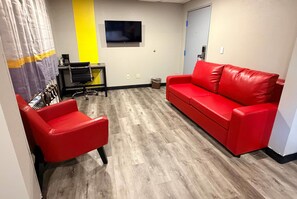 A room with a red sofa, a red chair, a TV on the wall, a desk with a chair, and a door.