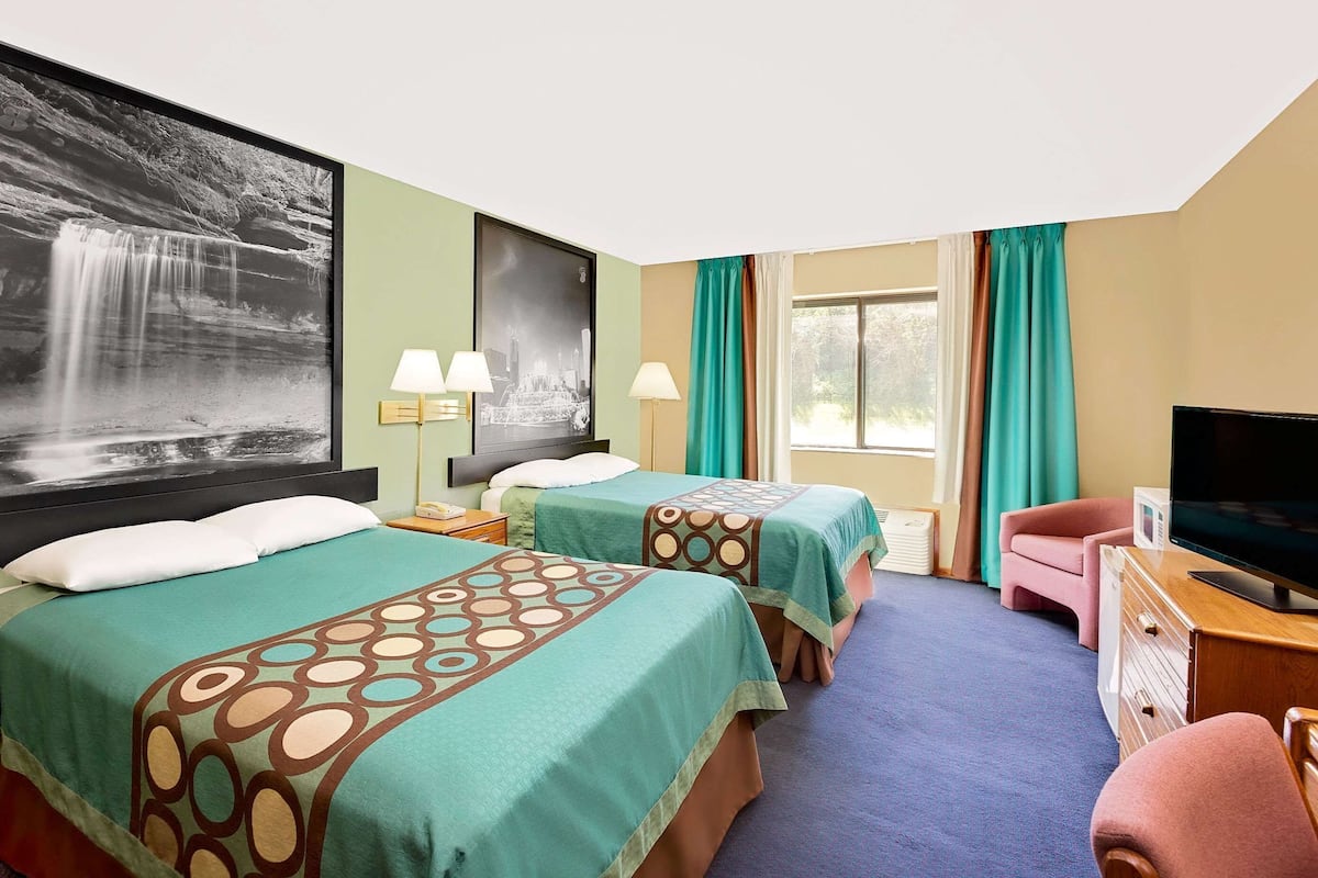 Quarto de hotel com duas camas, uma escrivaninha com televisão e uma janela grande com cortinas.