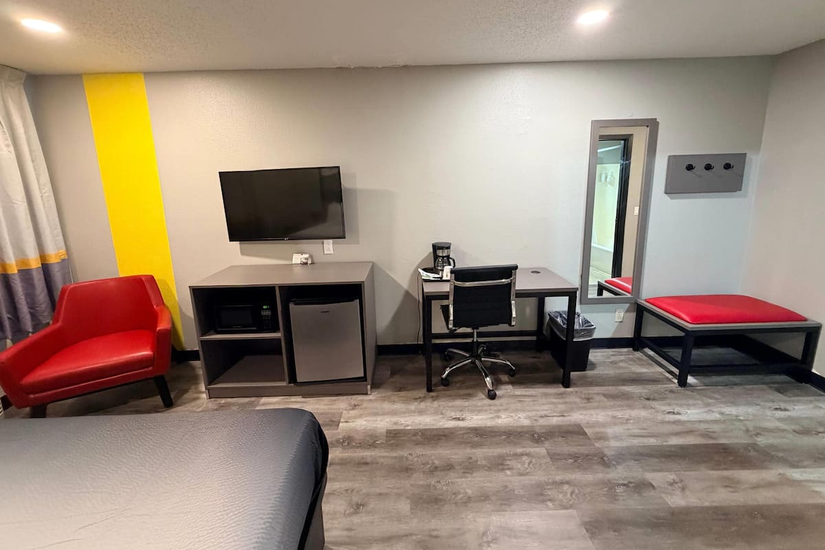 Quarto de hotel com cama, escrivaninha com computador, televisão, cadeira e um banco vermelho.