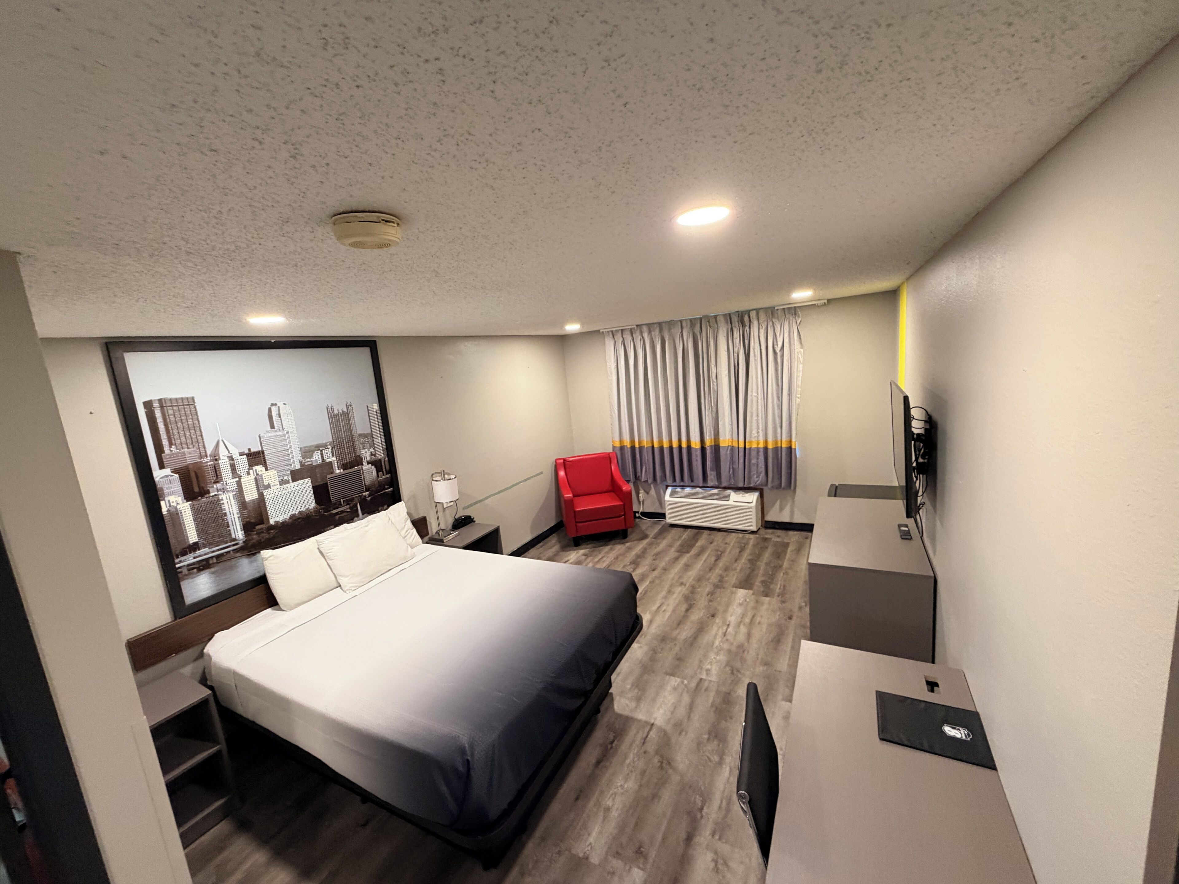 A hotel room with a bed, a red chair, a TV, and a large window with curtains.