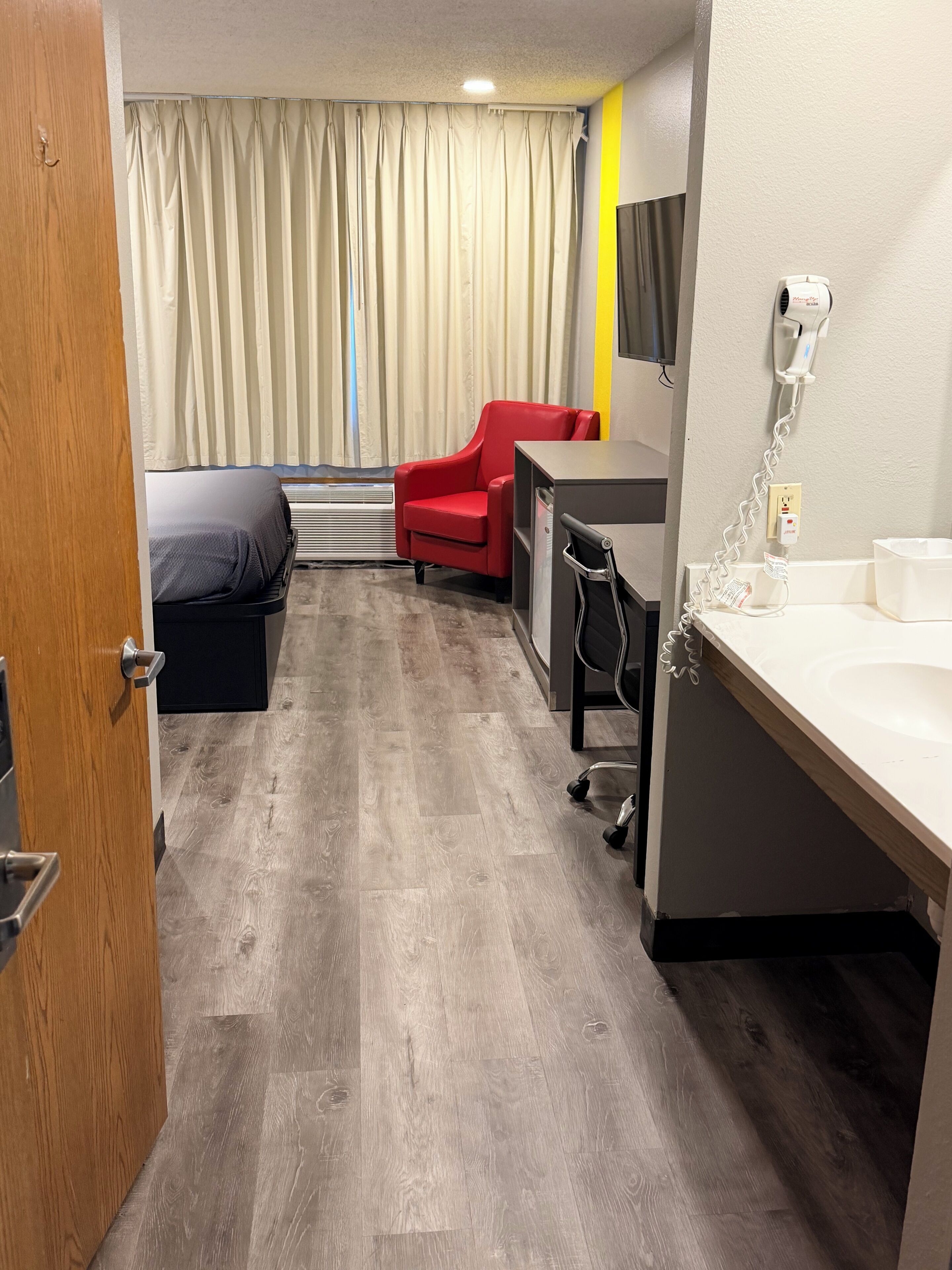 Ein Hotelzimmer mit Bett, Schreibtisch, rotem Sessel, Dusche und Vorhang.