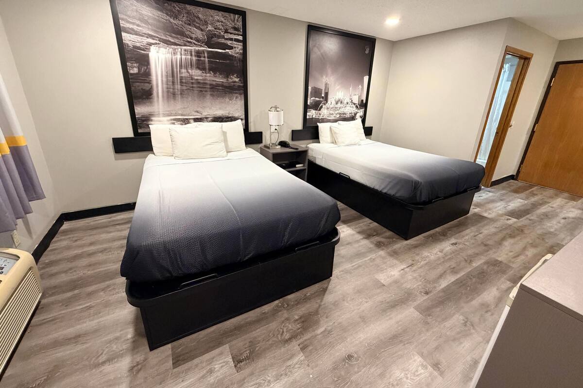 Quarto de hotel com duas camas, uma mesa de cabeceira, uma luminária e uma janela grande.