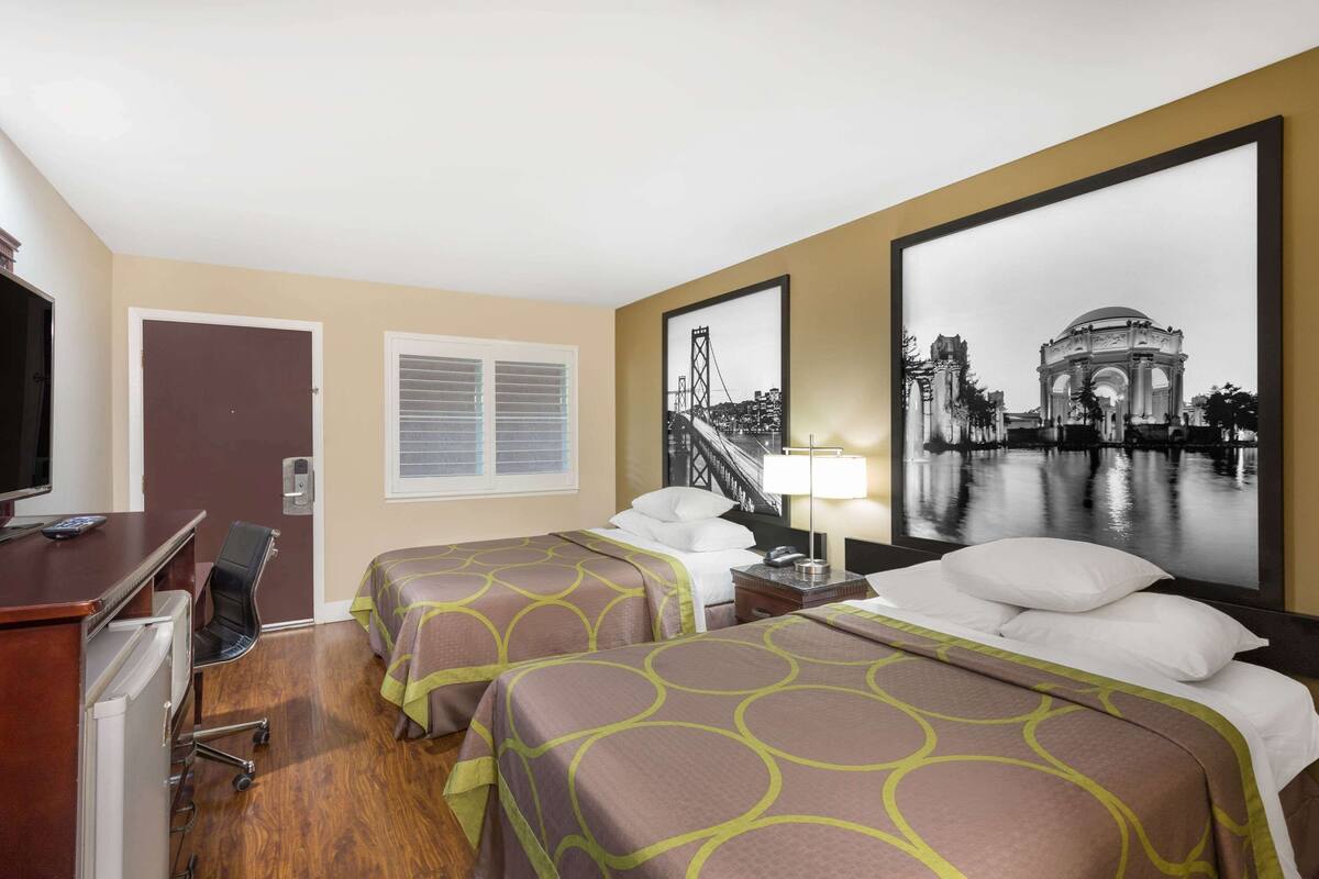 A hotel room with two beds, a desk with a computer, a TV, and a large framed picture of a bridge.