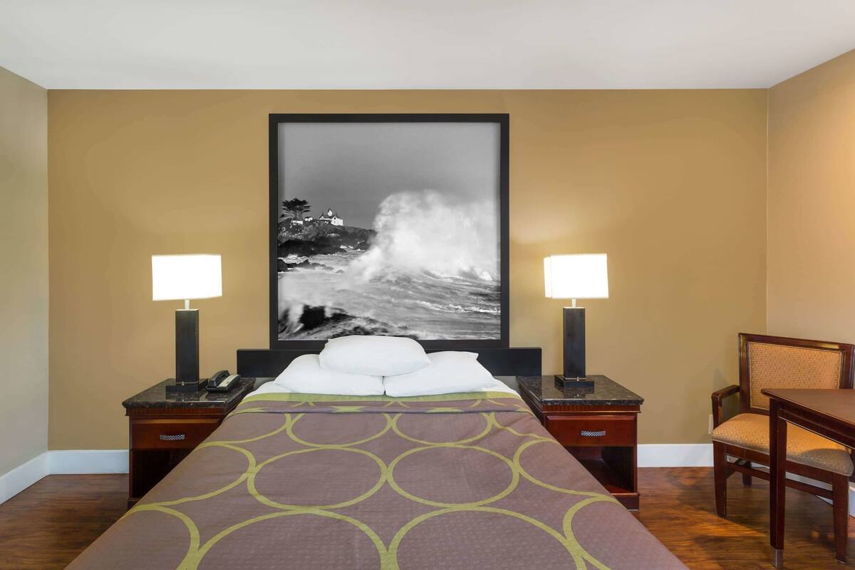 A hotel room with a bed, two bedside tables, a desk, a chair, and a large framed photograph of a wave crashing on a beach.