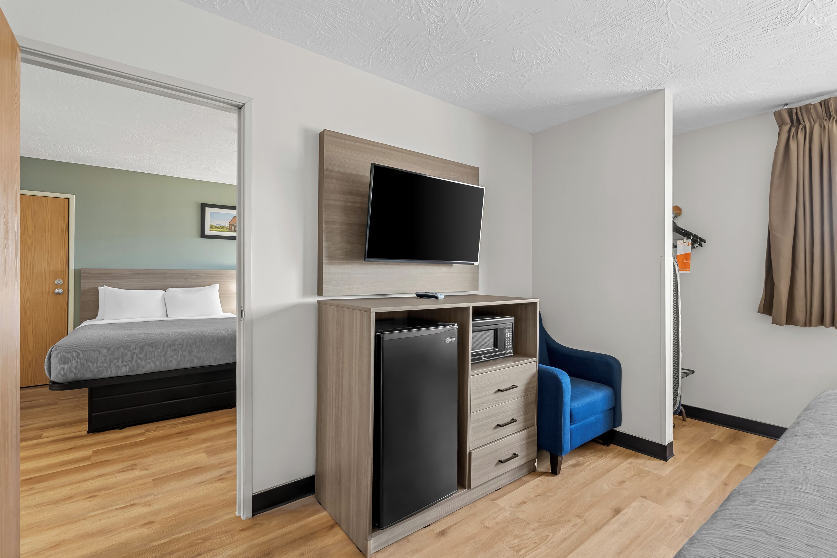 A modern hotel room with a bed, a desk with a flat-screen TV, a microwave, and a blue chair.