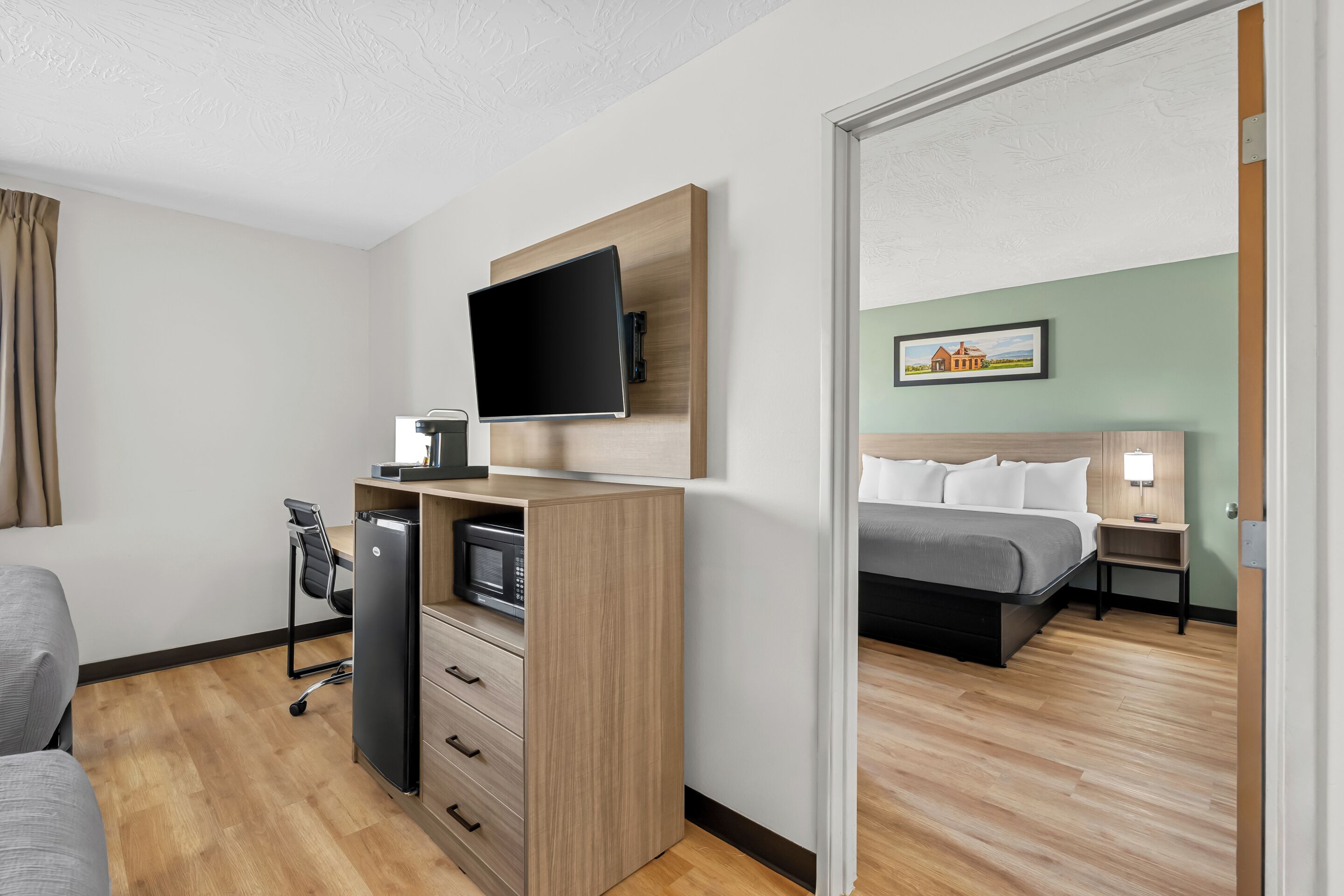 Ett hotellrum med en platt-TV som är monterad på ett träskåp, ett skrivbord med en dator och en mikrovågsugn, en säng med ett grått överkast och ett nattduksbord med en lampa.