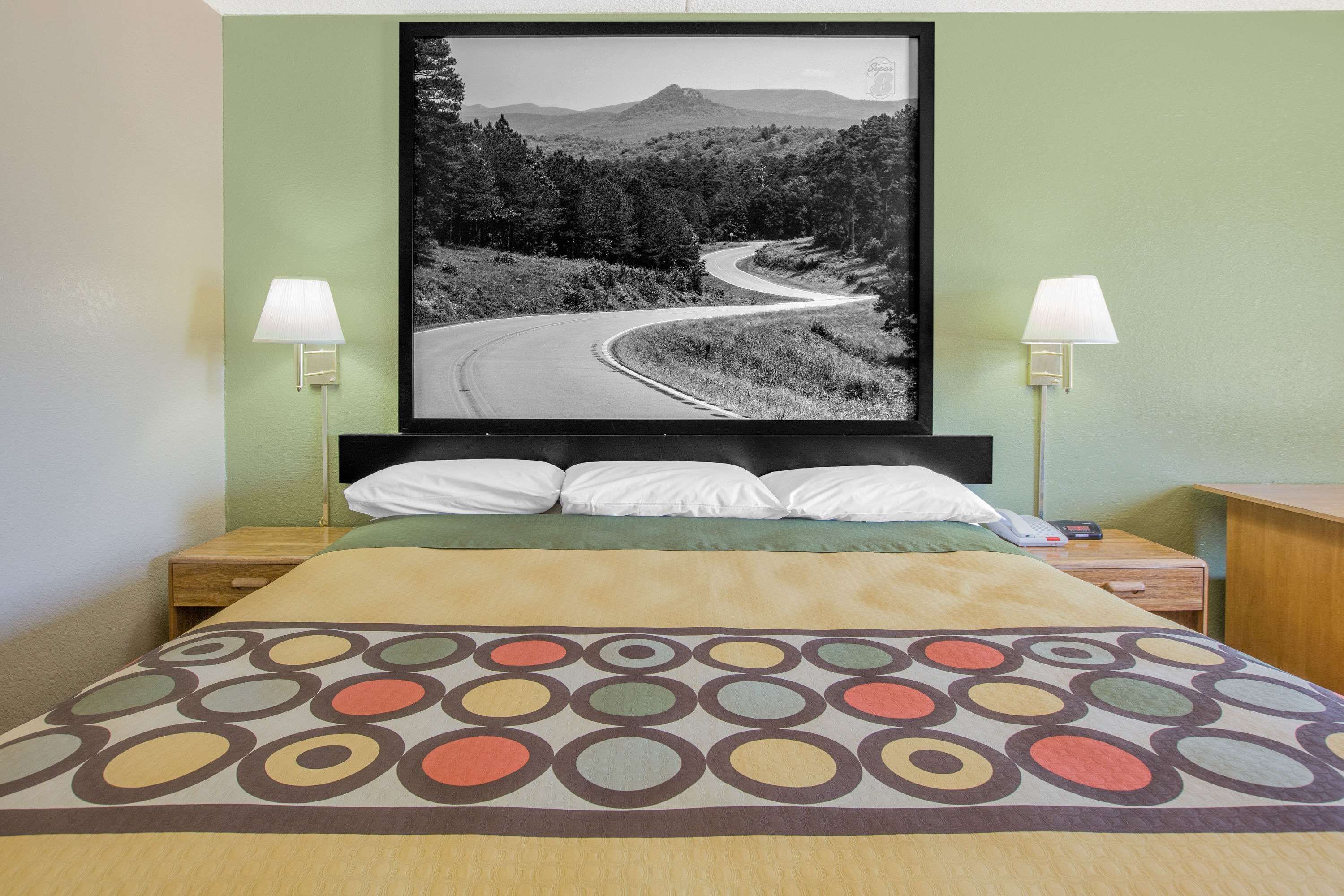 Una habitación de hotel con una cama, dos mesitas de noche, una lámpara y un cuadro con una carretera sinuosa.