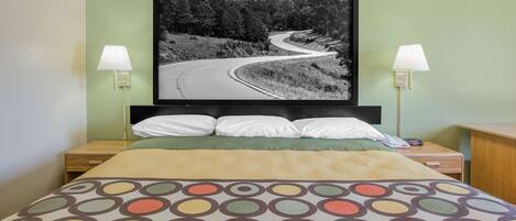 Una habitación de hotel con una cama, dos mesitas de noche, una lámpara y un cuadro con una carretera sinuosa.