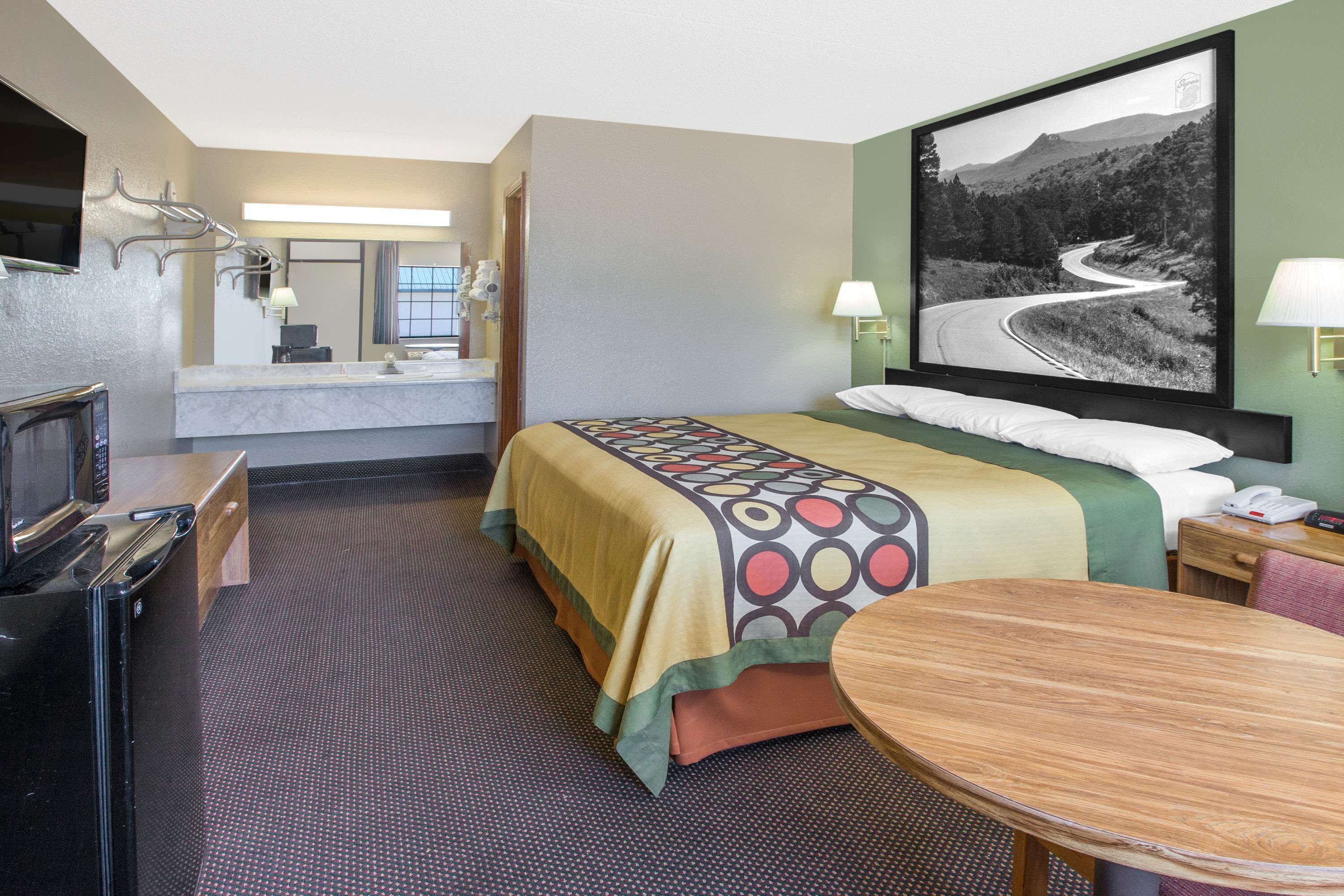 Ein Hotelzimmer mit einem Bett, einem Schreibtisch mit Computer, einem Fernseher, einem runden Holztisch und einem großen, gerahmten Bild an der Wand.