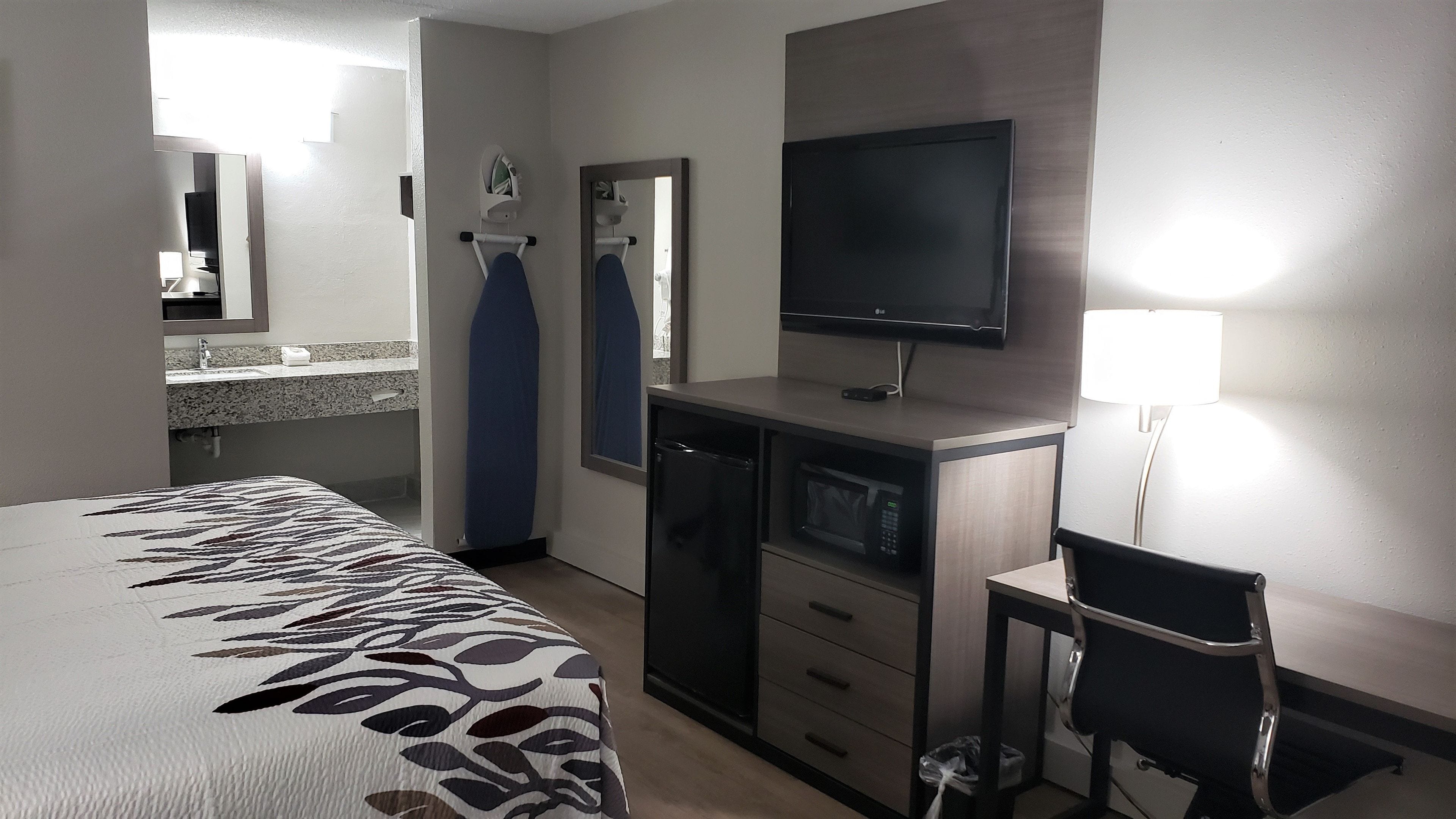 A hotel room with a bed, a desk with a television, a chair, a microwave, and a small trash bin.