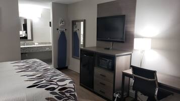 A hotel room with a bed, a desk with a television, a chair, a microwave, and a small trash bin.