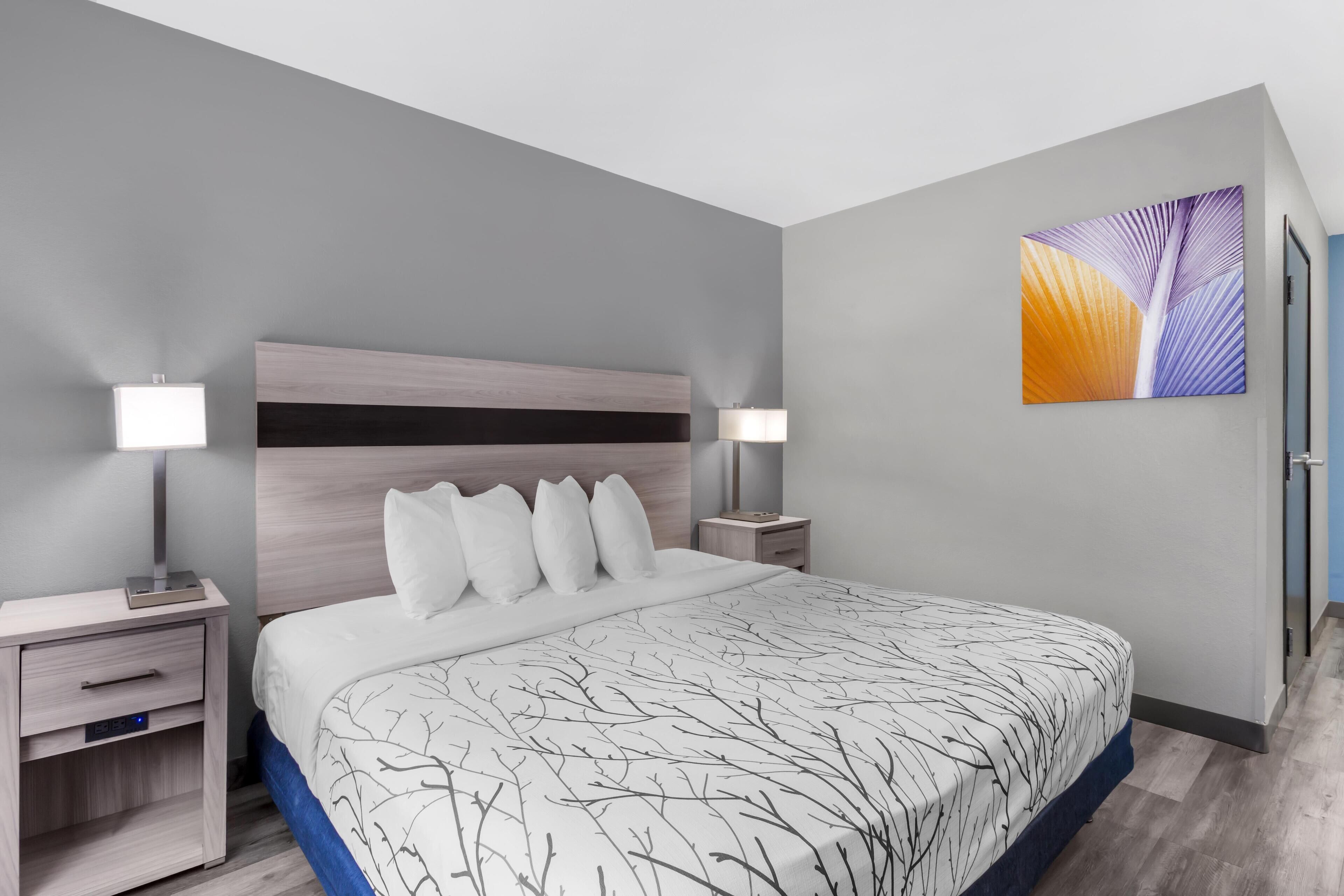 A modern hotel room with a large bed, two bedside tables, a nightstand, and a wall-mounted artwork.