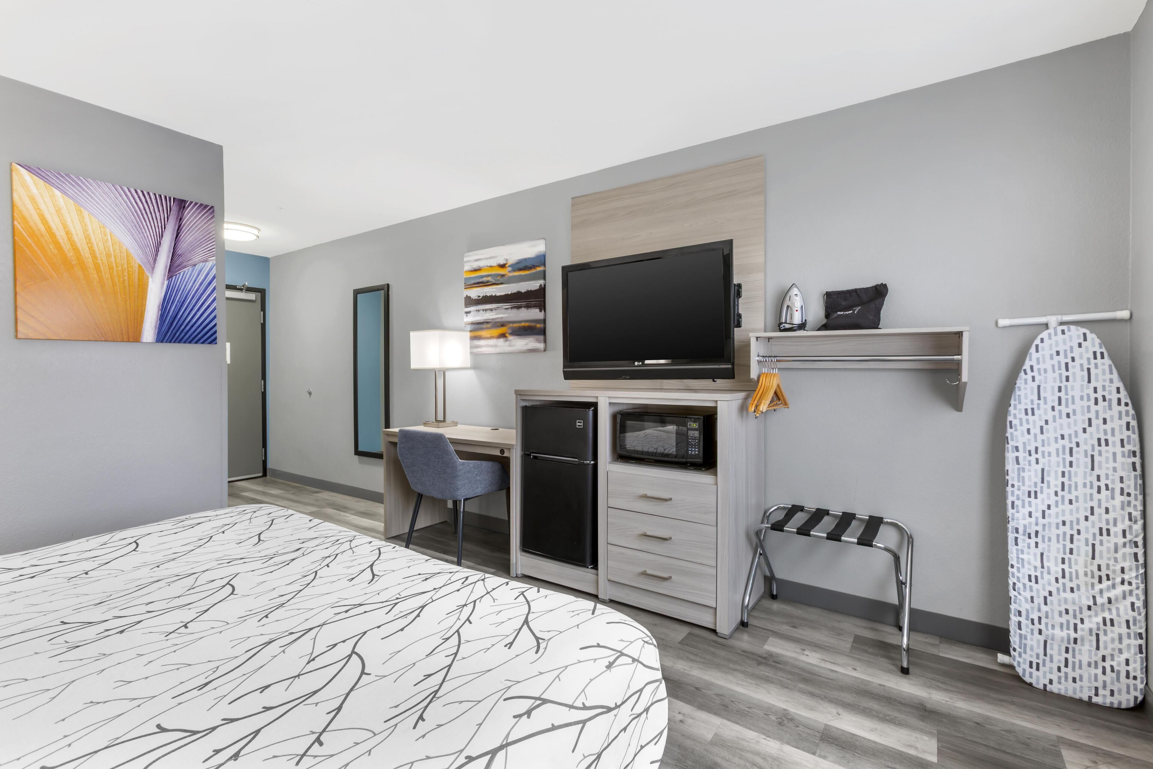 A hotel room with a bed, a desk with a computer, a TV, a microwave, and an ironing board.