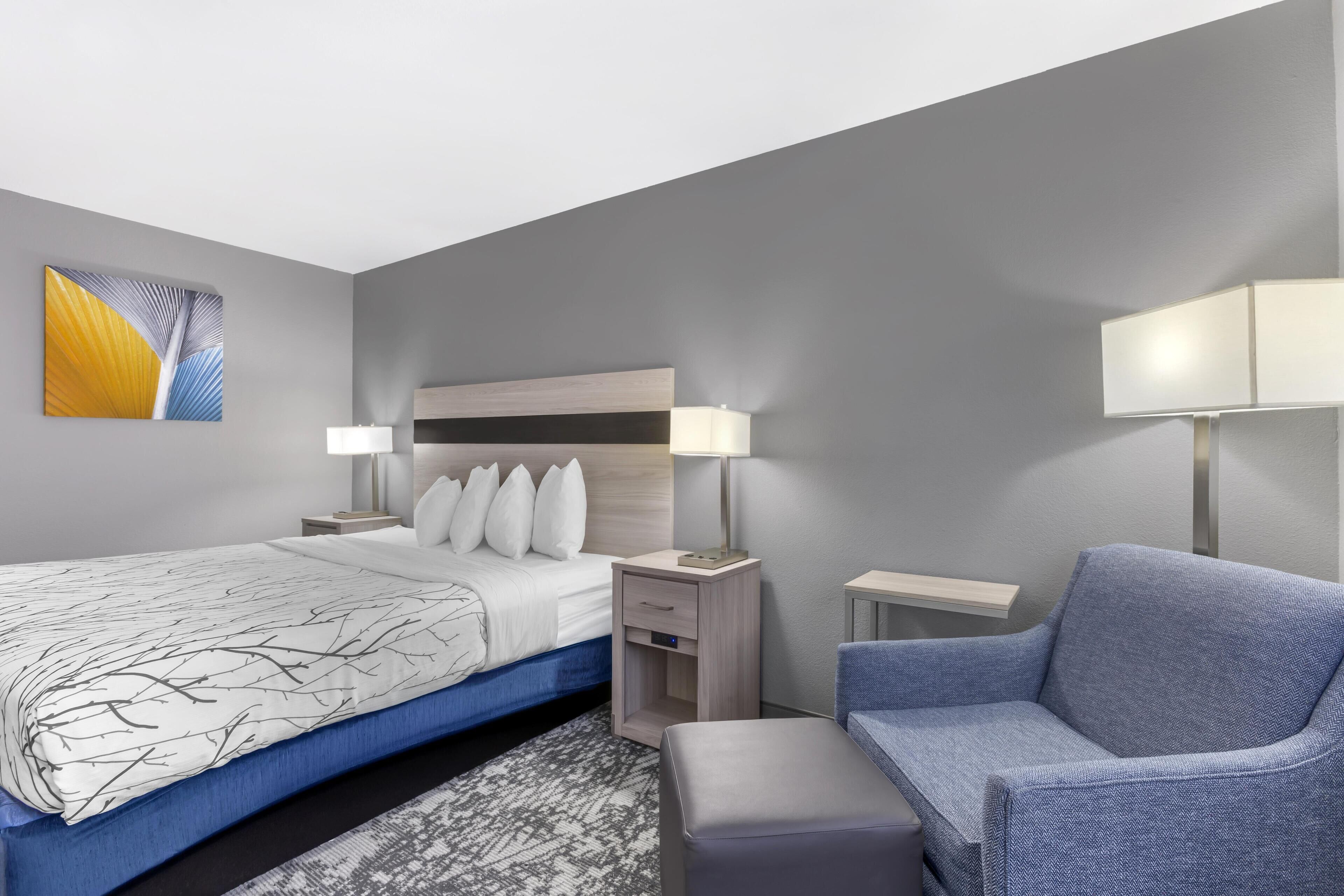 A modern hotel room with a large bed, two bedside tables with lamps, a blue armchair, and a small side table.