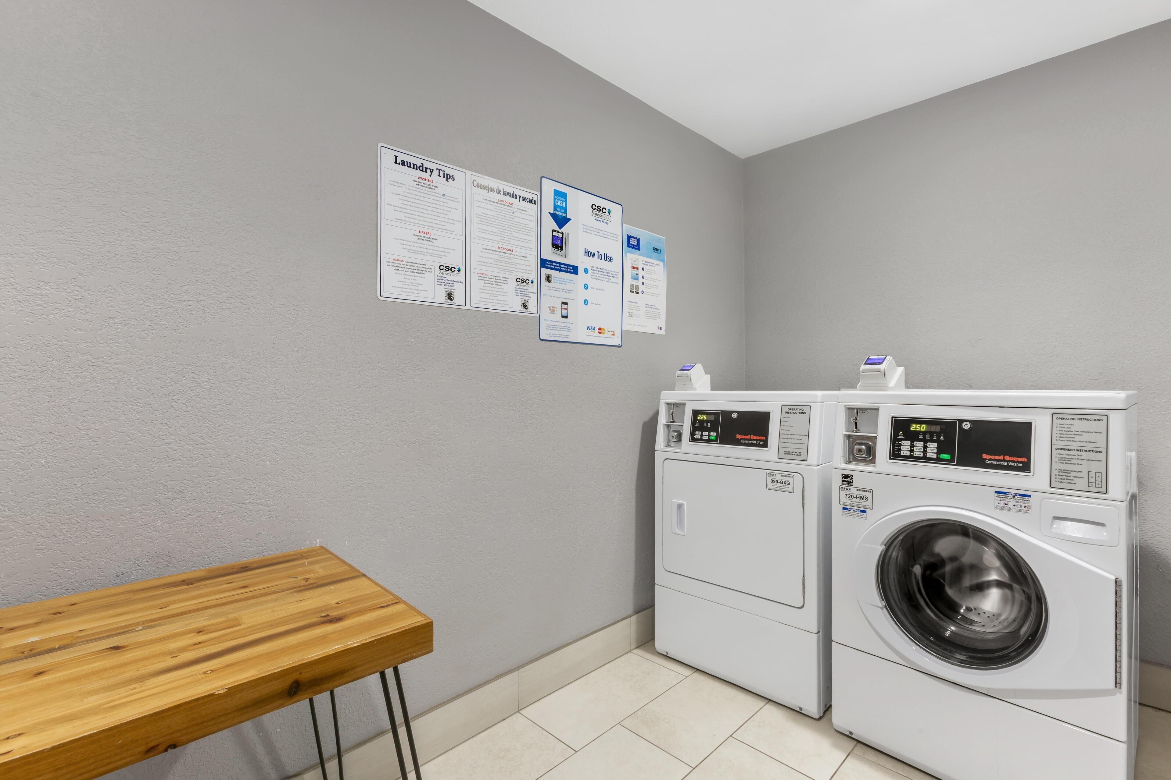 A laundry room with two washing machines, a wooden table, and informational posters on the wall.