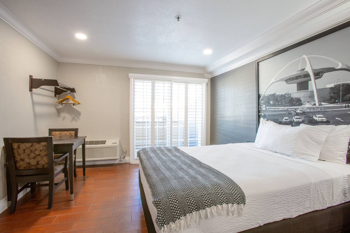A hotel room with a bed, a desk, and a chair. There is a window with blinds and a view of a bridge.