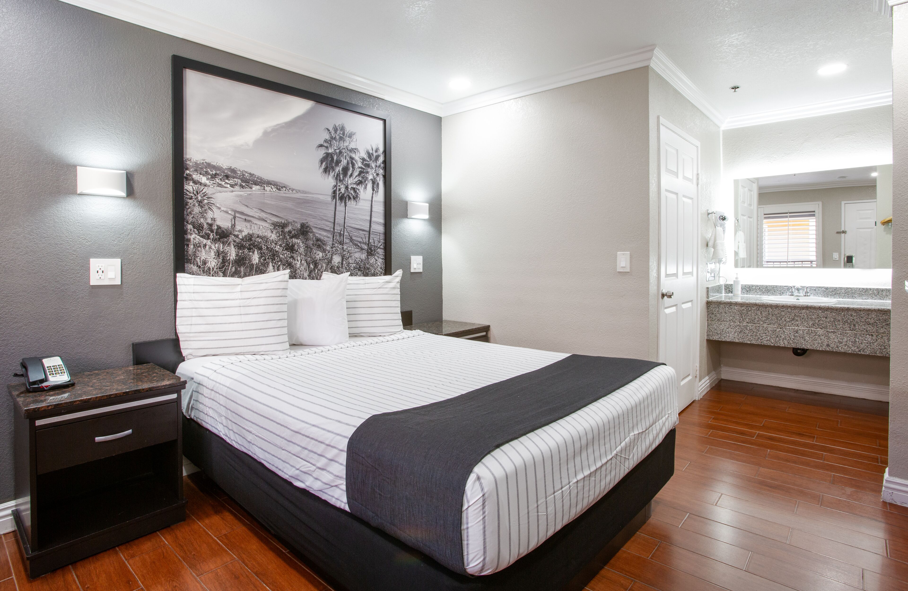 A neatly arranged bedroom with a large bed, bedside tables, a nightstand, a framed picture on the wall, and a bathroom visible through an open door.