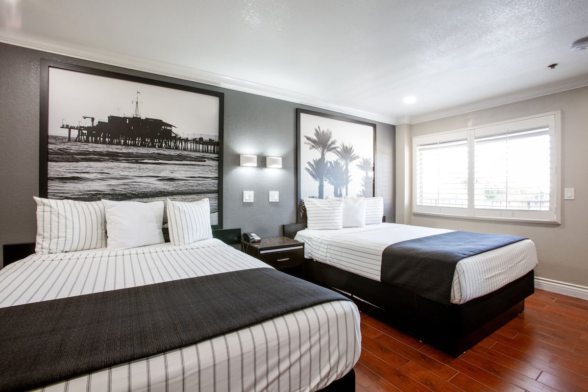 A hotel room with two beds, a large framed picture of a pier, a nightstand, and a window with blinds.