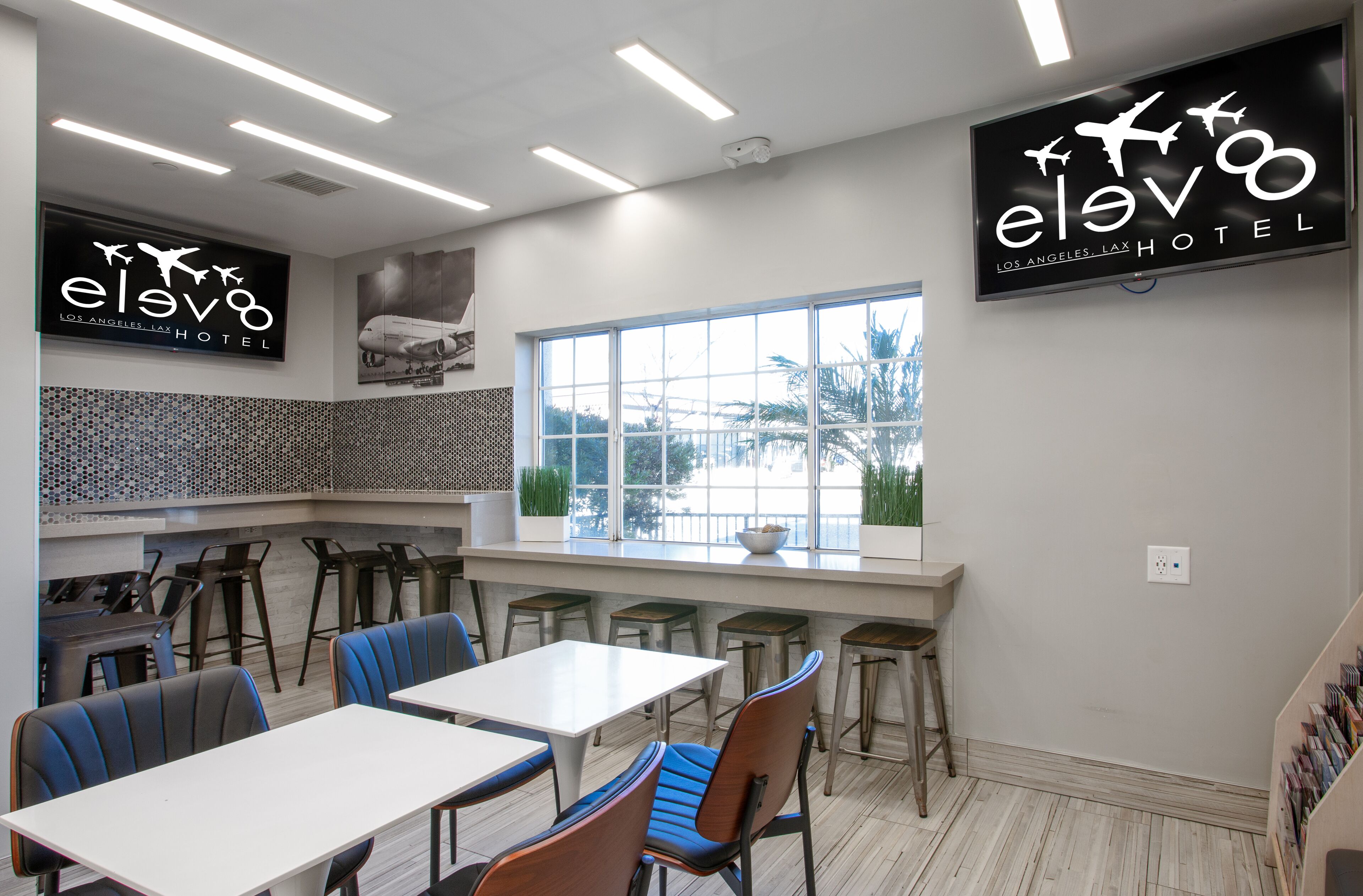 Ett modernt matområde med höga bord och stolar, ett stort fönster med utsikt och en väggmonterad skylt för hotellet Elvo Los Angeles LAX.