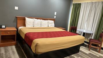Una habitación de hotel con cama, mesita de noche, silla, pequeña mesa y ventana con cortinas.
