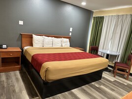 Una habitación de hotel con cama, mesita de noche, silla, pequeña mesa y ventana con cortinas.